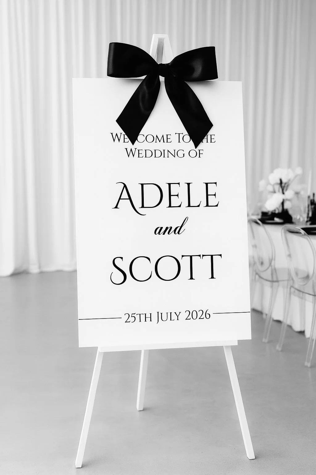 A wedding welcome sign with black text on a white background, decorated with a black ribbon bow at the top, mentioning Adele and Scott's wedding on July 25, 2026, with chairs and floral arrangements in the background.