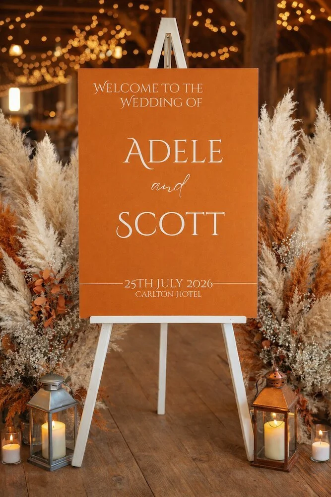 Orange wedding welcome sign surrounded by pampas grass and candles with fairy lights in the background.