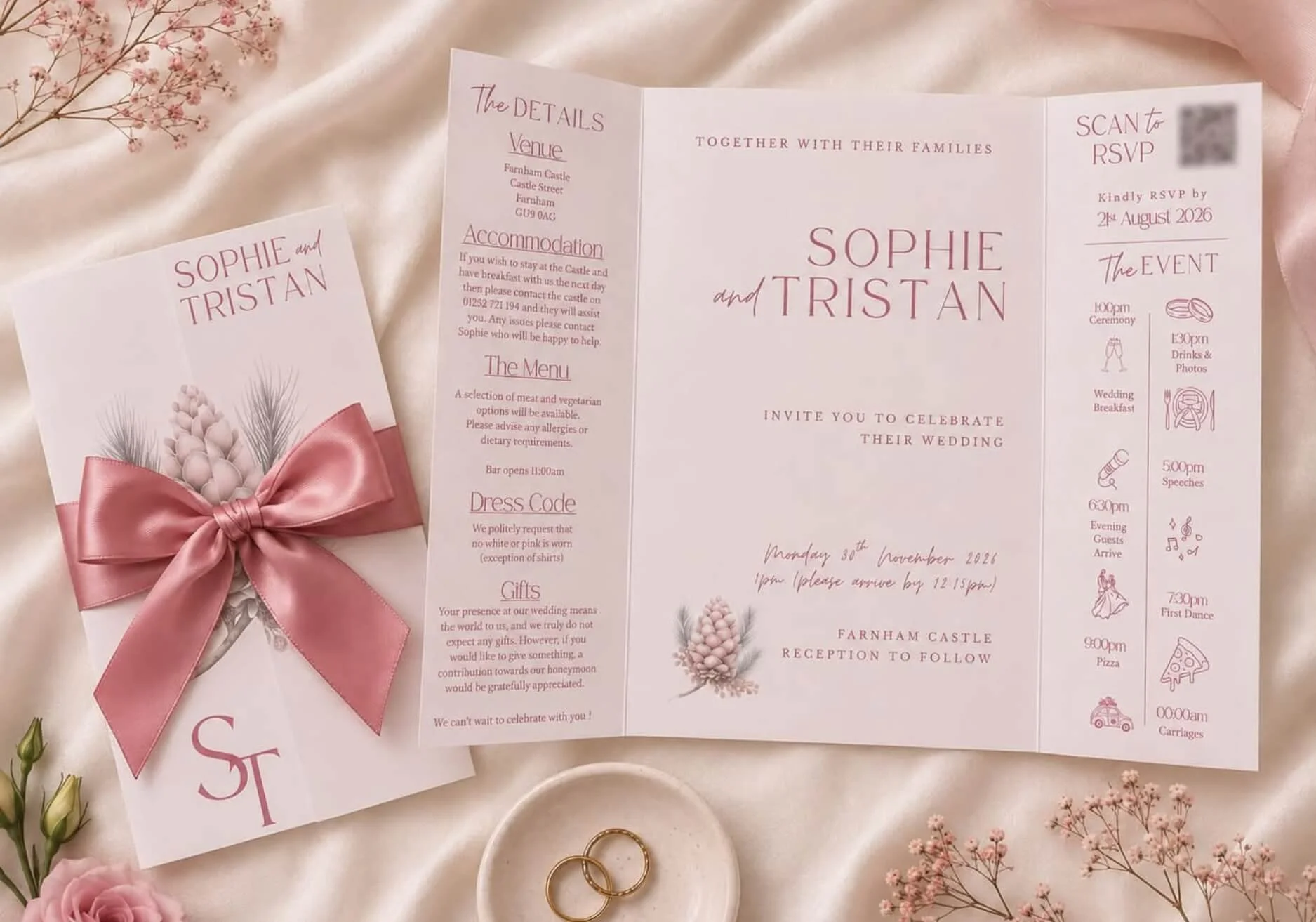 Wedding invitation leaflet and an RSVP card on cream-colored silk fabric. The invitation has pink text, a pink ribbon tied in a bow, and floral decorations. The RSVP card has a QR code. There are two gold rings in a small dish, pink roses, and sprigs of pink baby's breath flowers surrounding the cards.