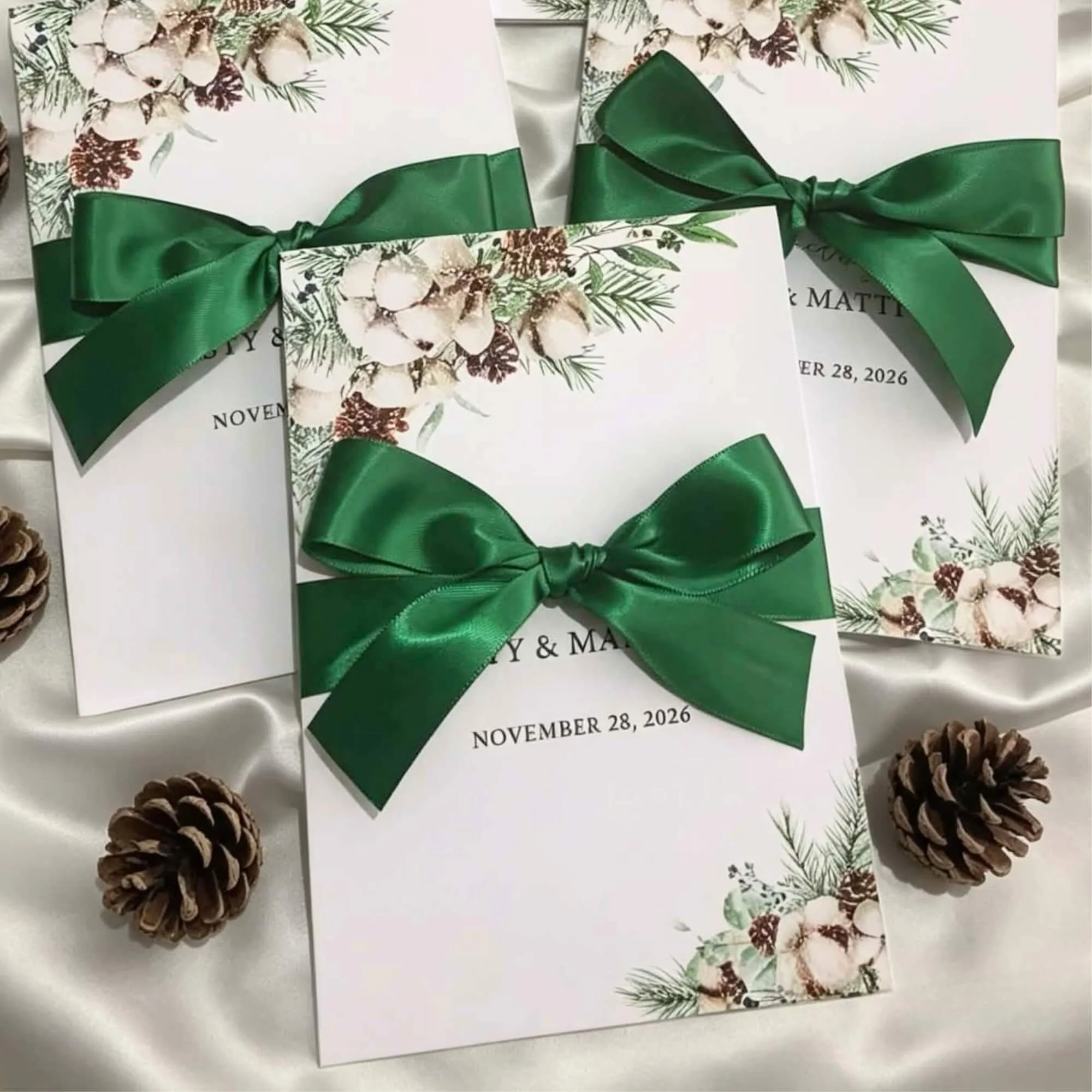 Wedding invitations with a winter theme featuring pinecones, greenery, and white flowers, tied with a green satin ribbon and script including the date November 28, 2026.