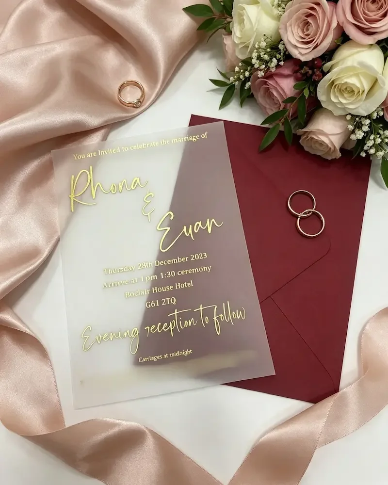 Wedding invitation card with gold text, a burgundy envelope, a gold ring, and a bouquet of pink and white roses on a satin fabric.