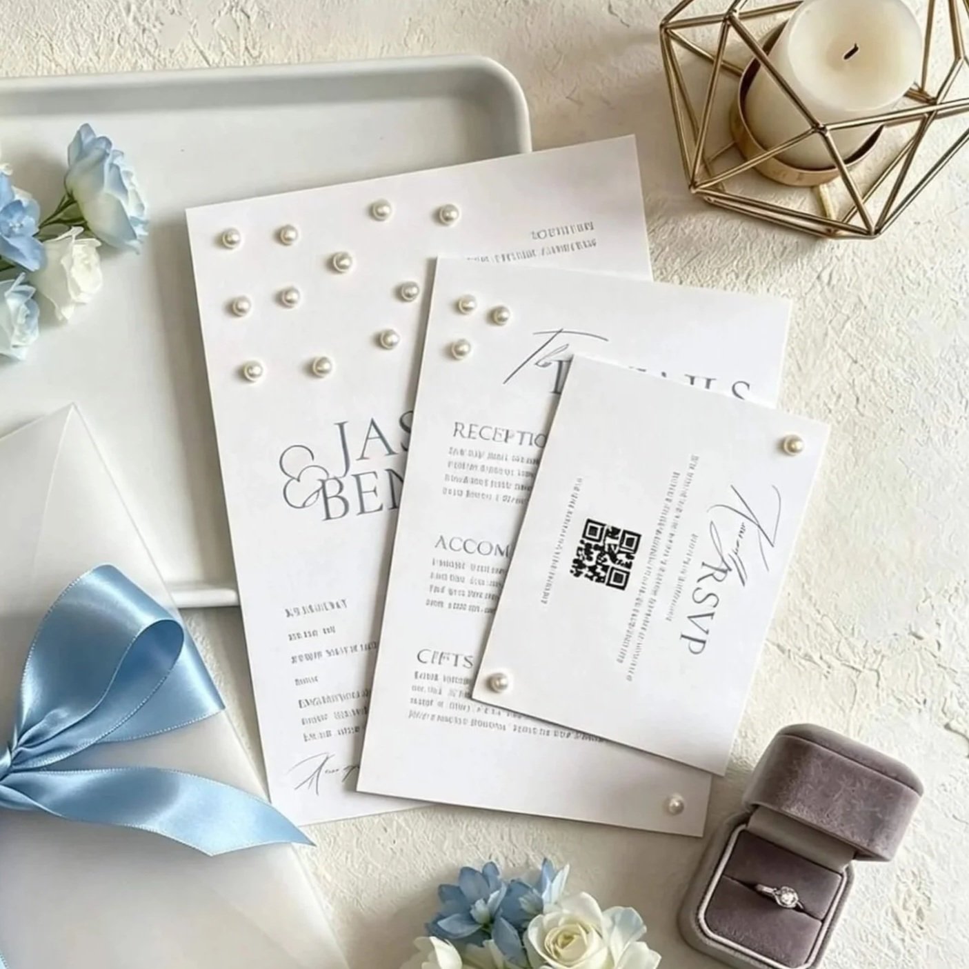 Elegant wedding invitation cards with pearl embellishments, a light blue satin ribbon, a velvet ring box with a diamond ring, light blue and white flowers, and a candle in a decorative holder on a white textured surface.