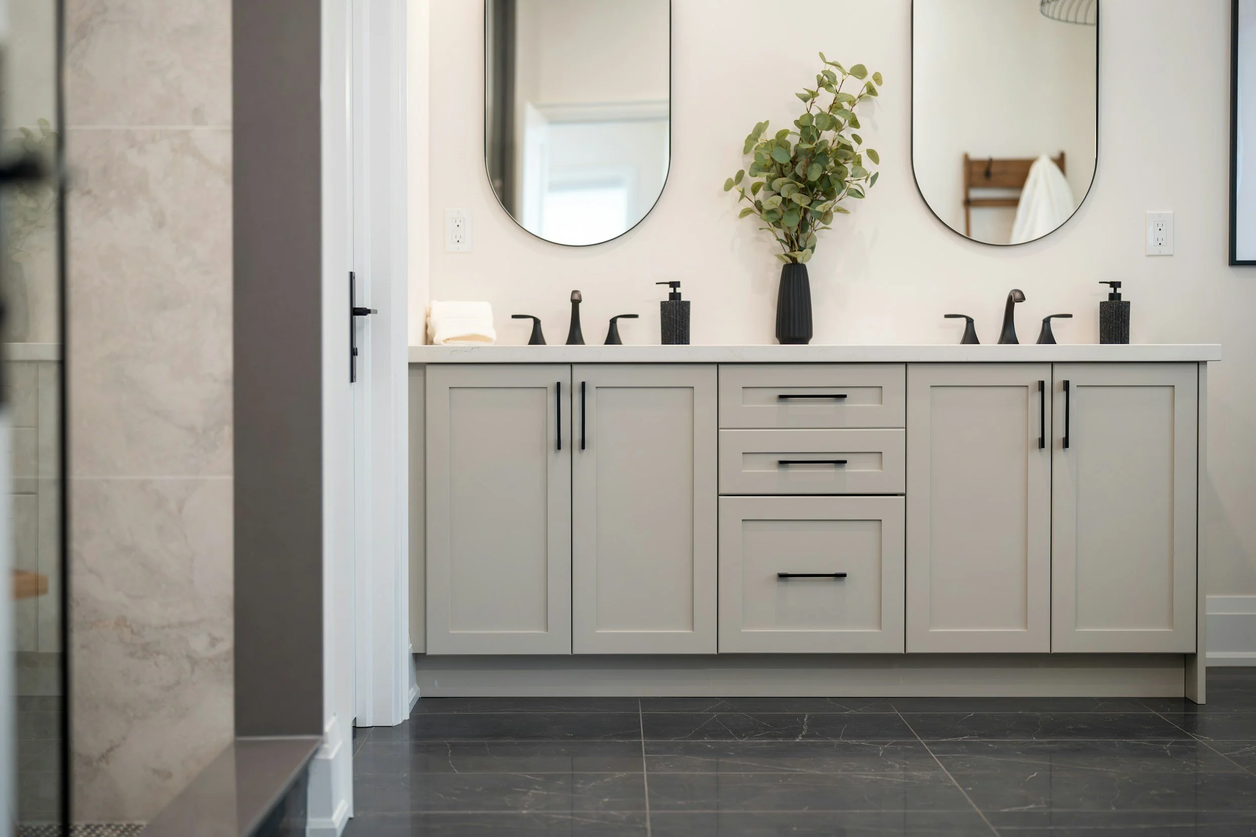 Modern bathroom vanity with two oval mirrors, black faucets, a black soap dispenser, a potted plant, and towels on a white countertop against a white wall.