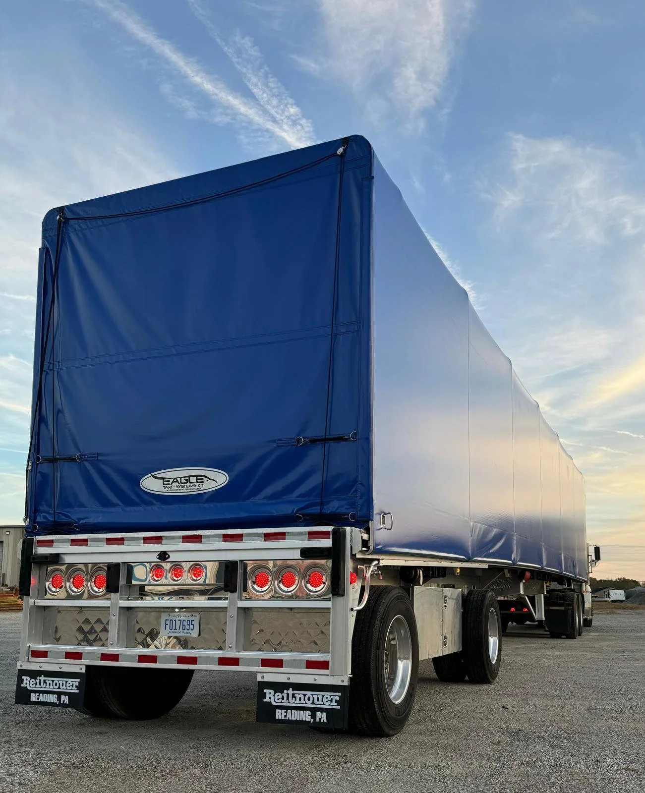 A large semi-truck with a blue covered trailer parked on an open lot under a sky with scattered clouds, sunset lighting, and a horizon with distant buildings.