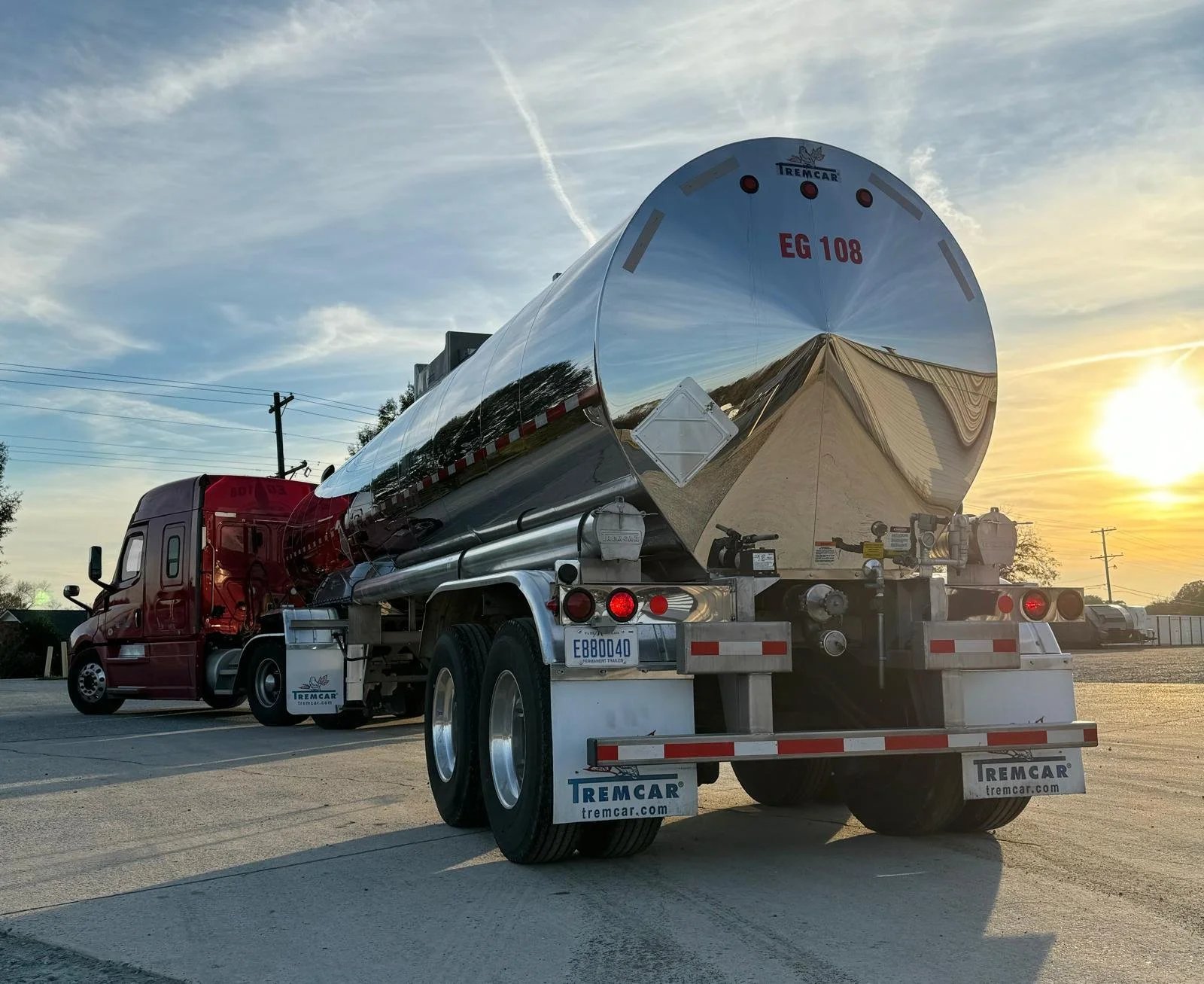 A shiny metal fuel tanker truck parked on a concrete lot during sunset with power lines and trees in the background.