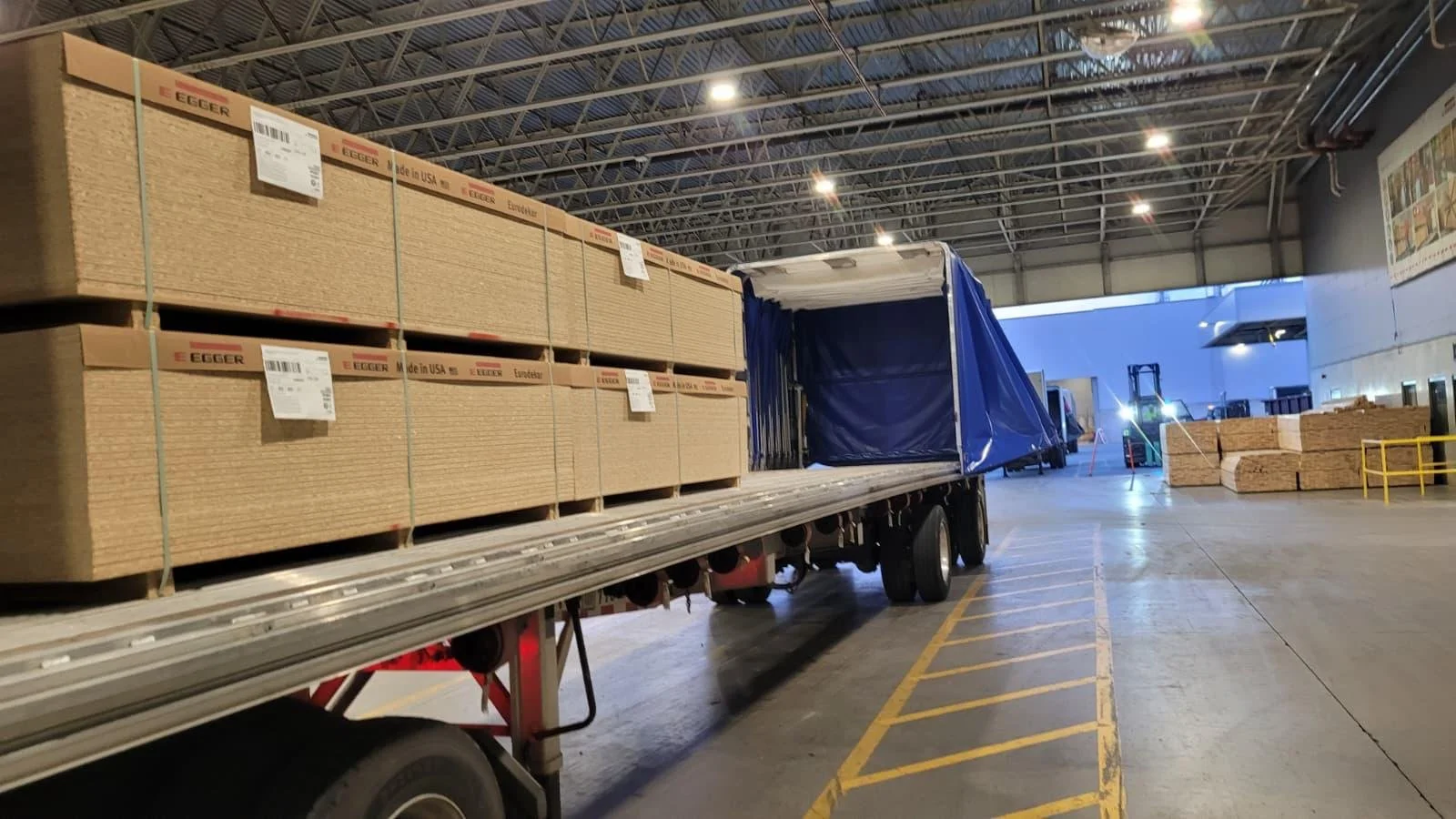 Inside a warehouse, a flatbed truck loaded with large cardboard boxes is parked. The boxes are labeled "Egger" and marked as made in the USA. The warehouse has high ceilings with bright overhead lighting, and additional stacks of cardboard are visible in the background.