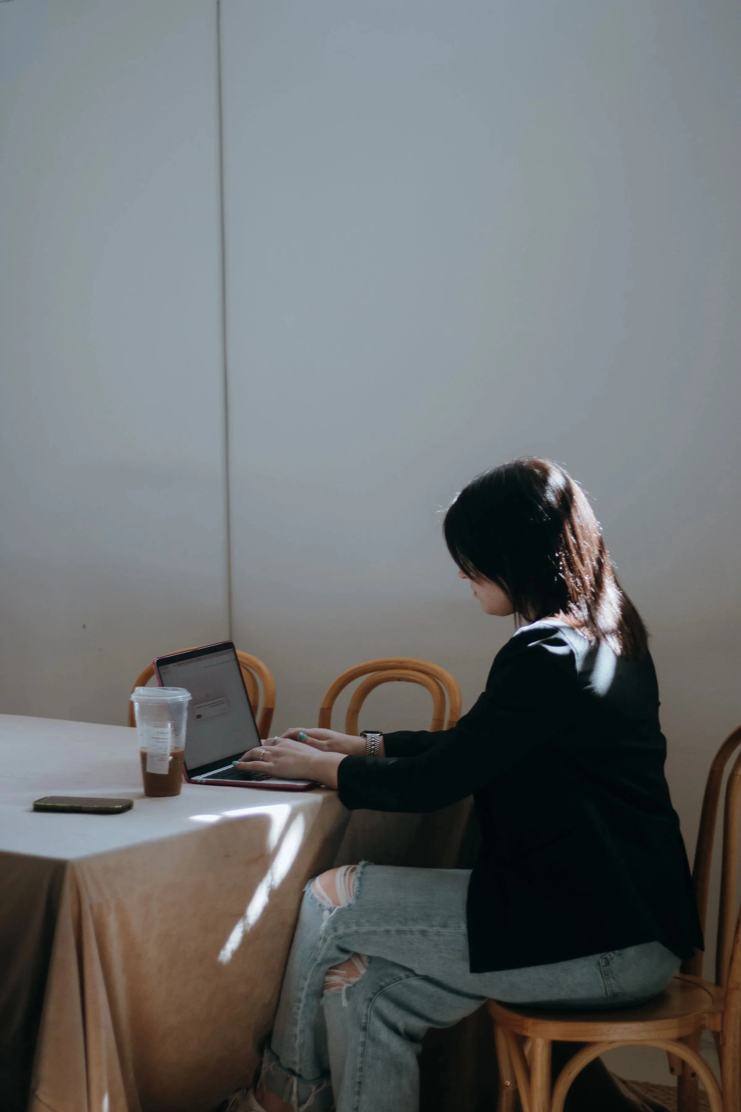 A woman sitting at a table working on a laptop, with a Starbucks drink and phone on the table, in a room with plain walls and wooden chairs.