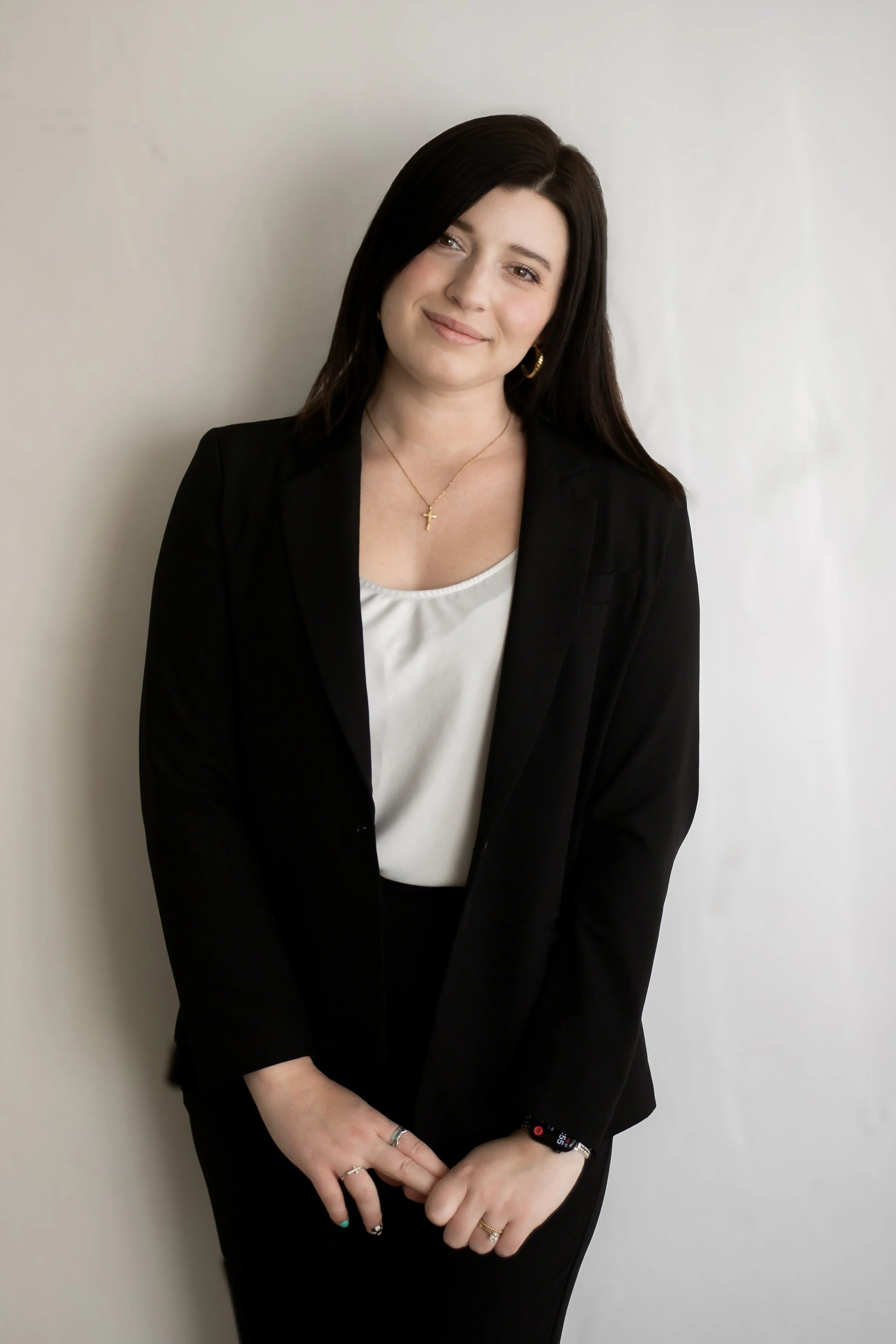 A woman with dark brown hair, wearing a black blazer and a white top, standing against a plain light-colored wall. She is smiling slightly, with her hands clasped in front of her, wearing jewelry including rings, a necklace with a cross, earrings, and a smartwatch.