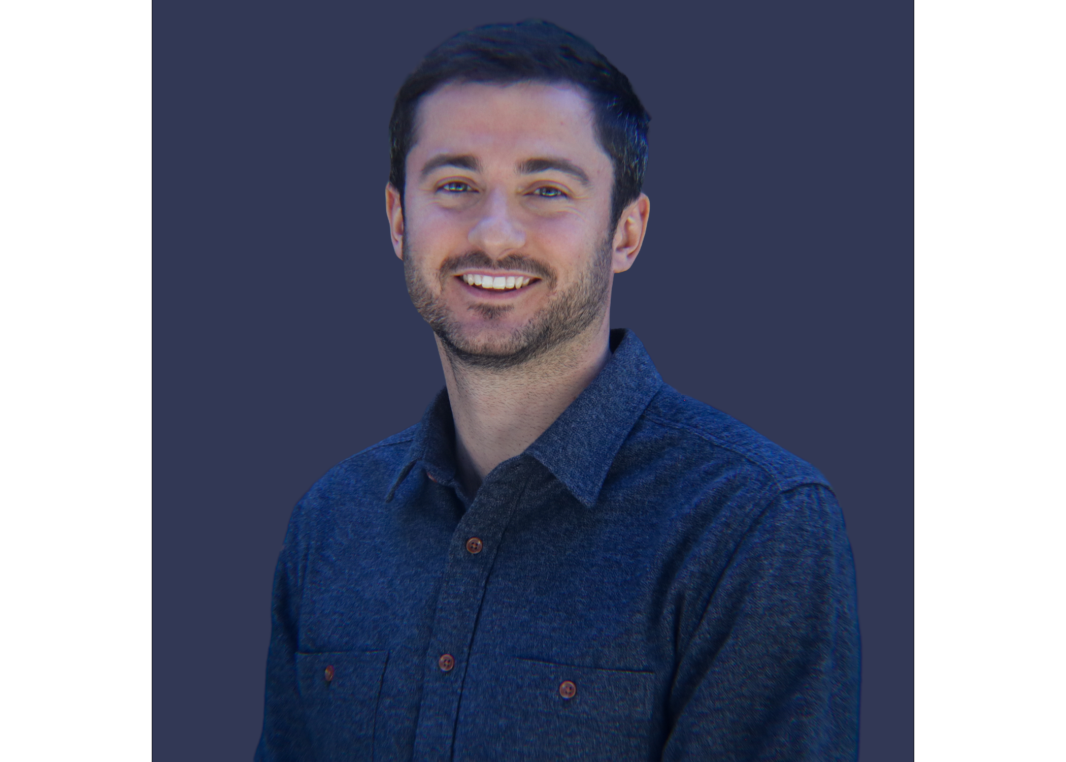 A young man with short dark hair and a beard, wearing a blue button-up shirt, smiling with white teeth, against a dark background.