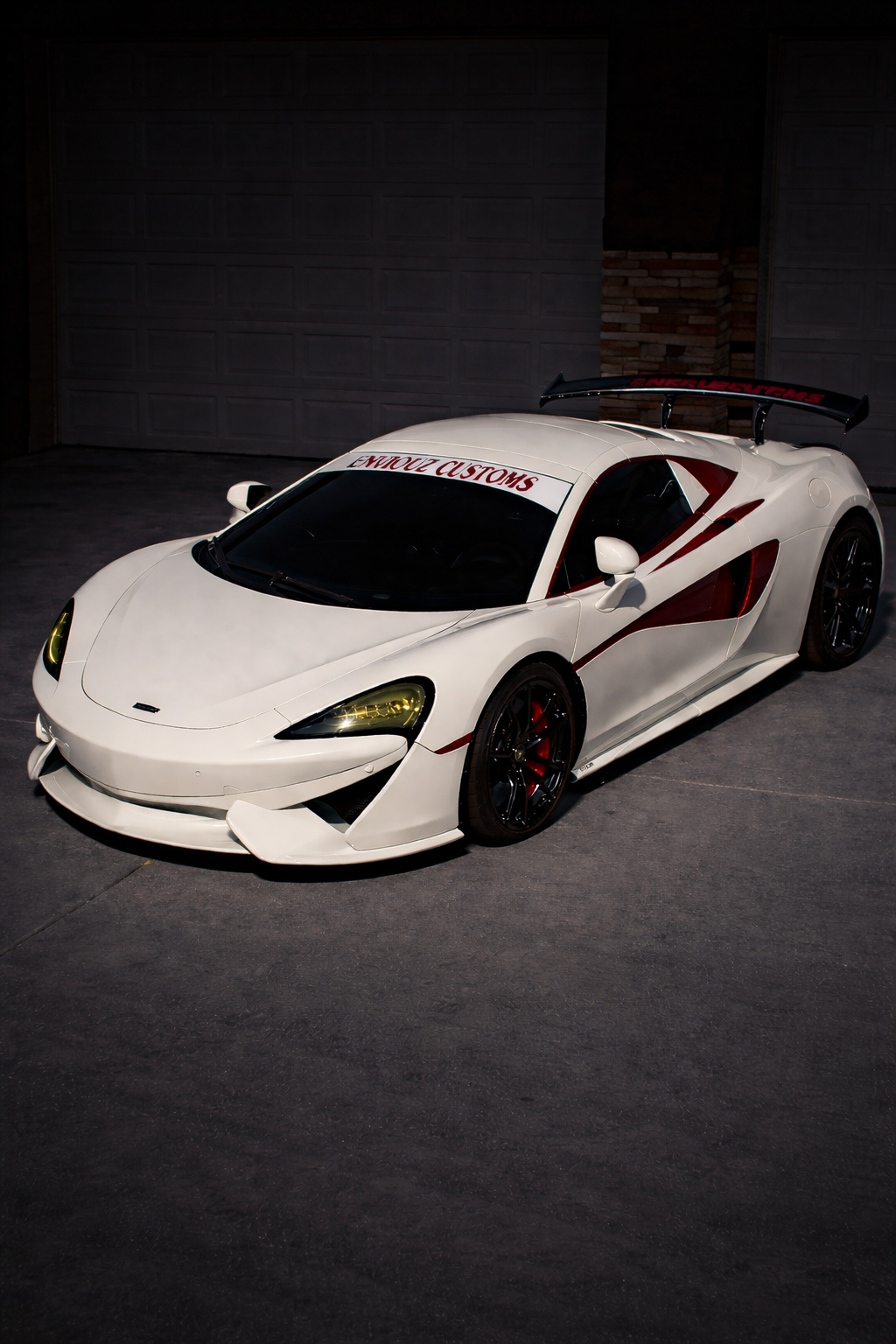 A white sports car parked in a driveway with a custom decal, black rims, and a large rear wing, under garage lighting.
