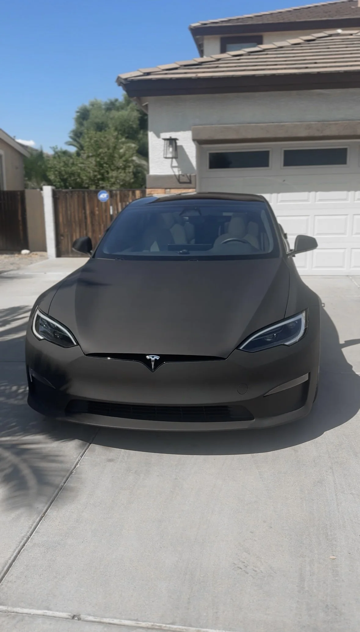Front view of a matte black Tesla electric car parked in a residential driveway in front of a white garage door and a house with a brown tiled roof.