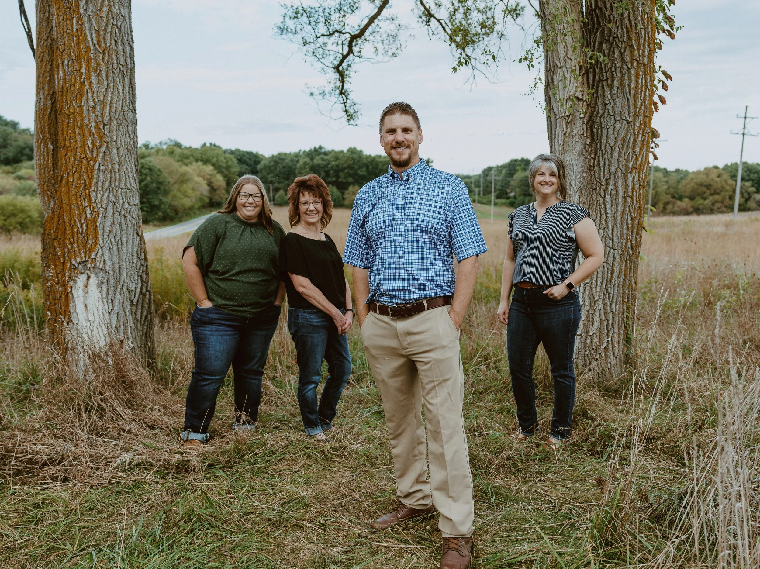 A group of four people, two women and two men, standing outdoors between two large trees with a scenic background of fields and trees, smiling at the camera.
