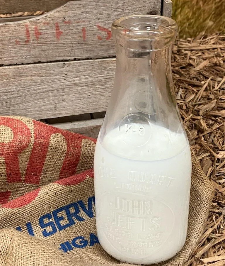 Glass bottle filled with milk, placed on a burlap sack with red and blue text, outdoors near a wooden crate and mulch.