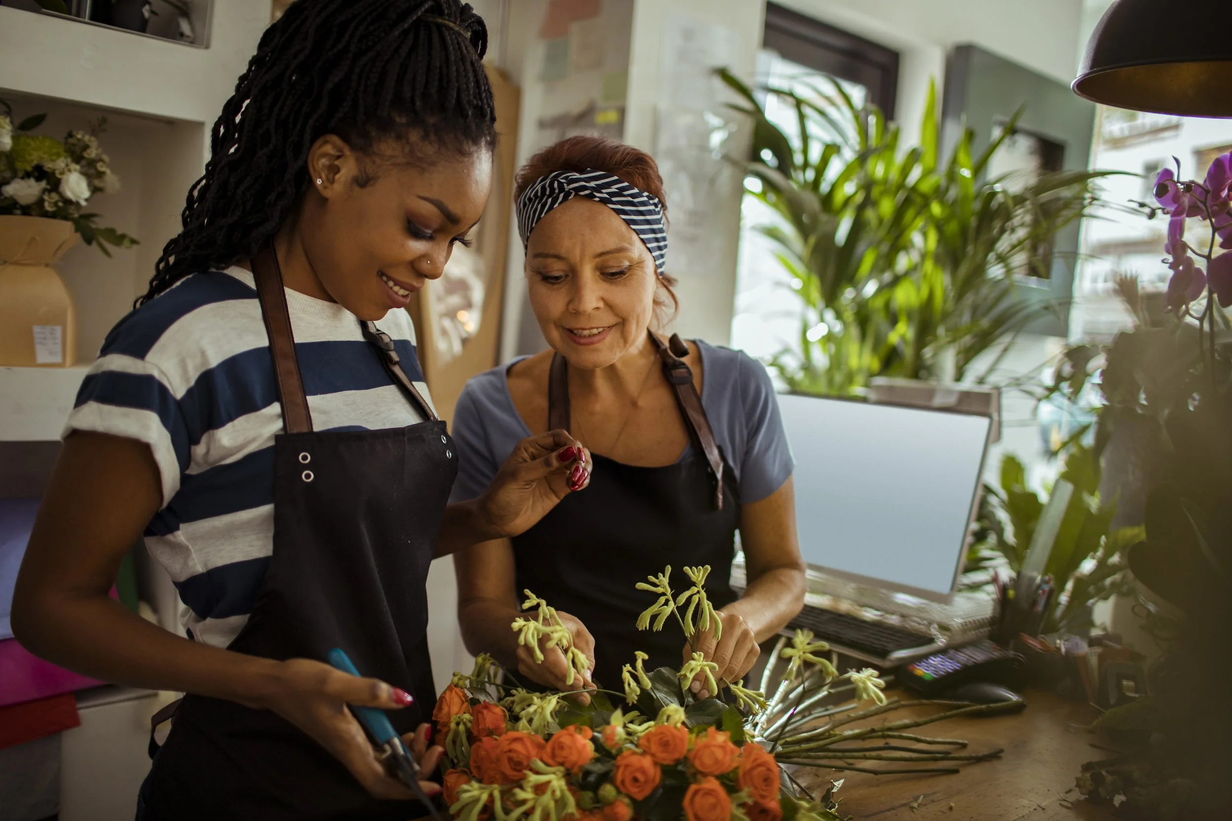 Two women, one younger with dark skin and dreadlocks and one older with light skin and short hair wearing a striped headscarf, are working together to arrange a floral display of orange roses and yellow flowers in a florist shop, surrounded by green plants and floral supplies.