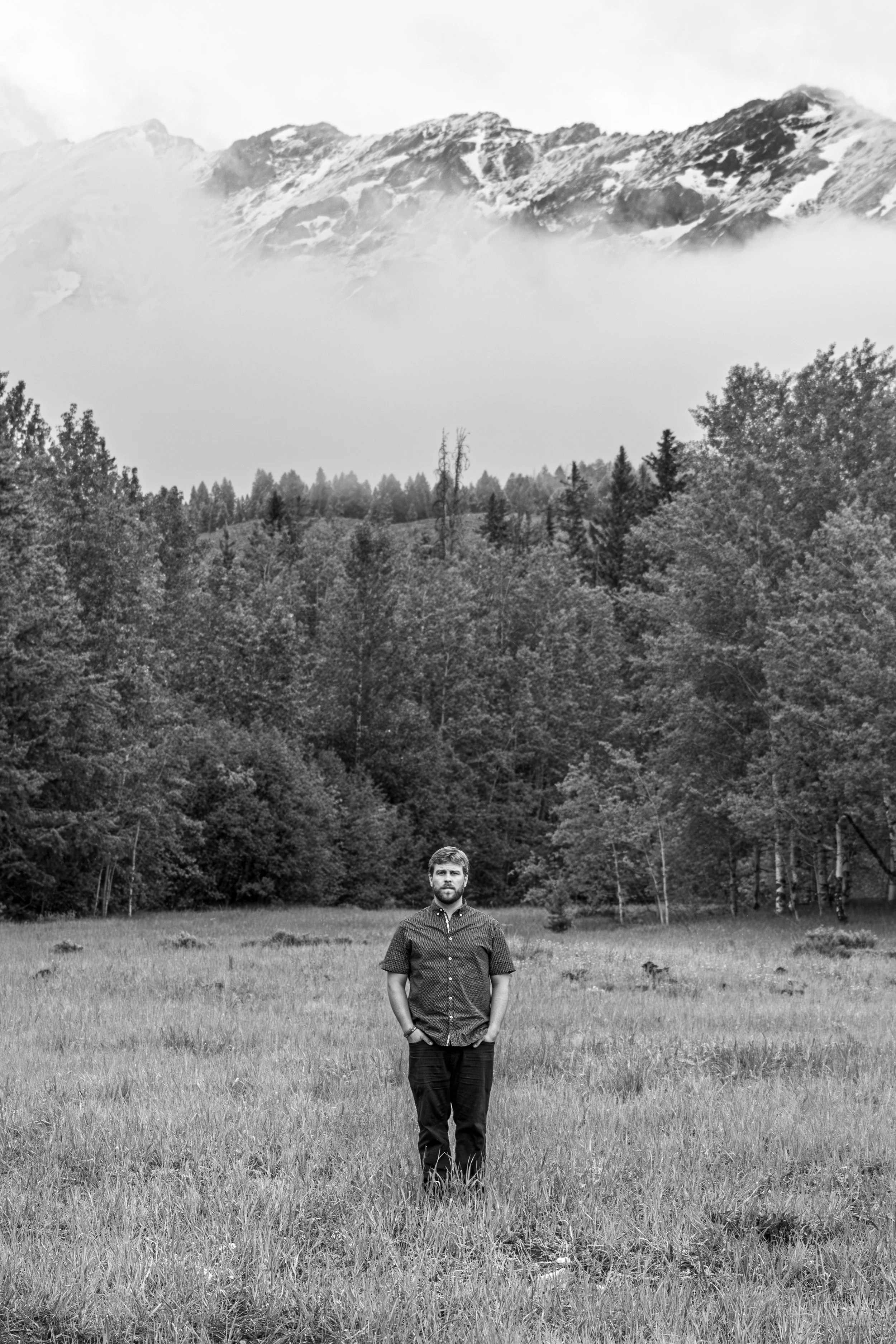 A man stands in a grassy field with trees behind him, mountains in the distance, in black and white.