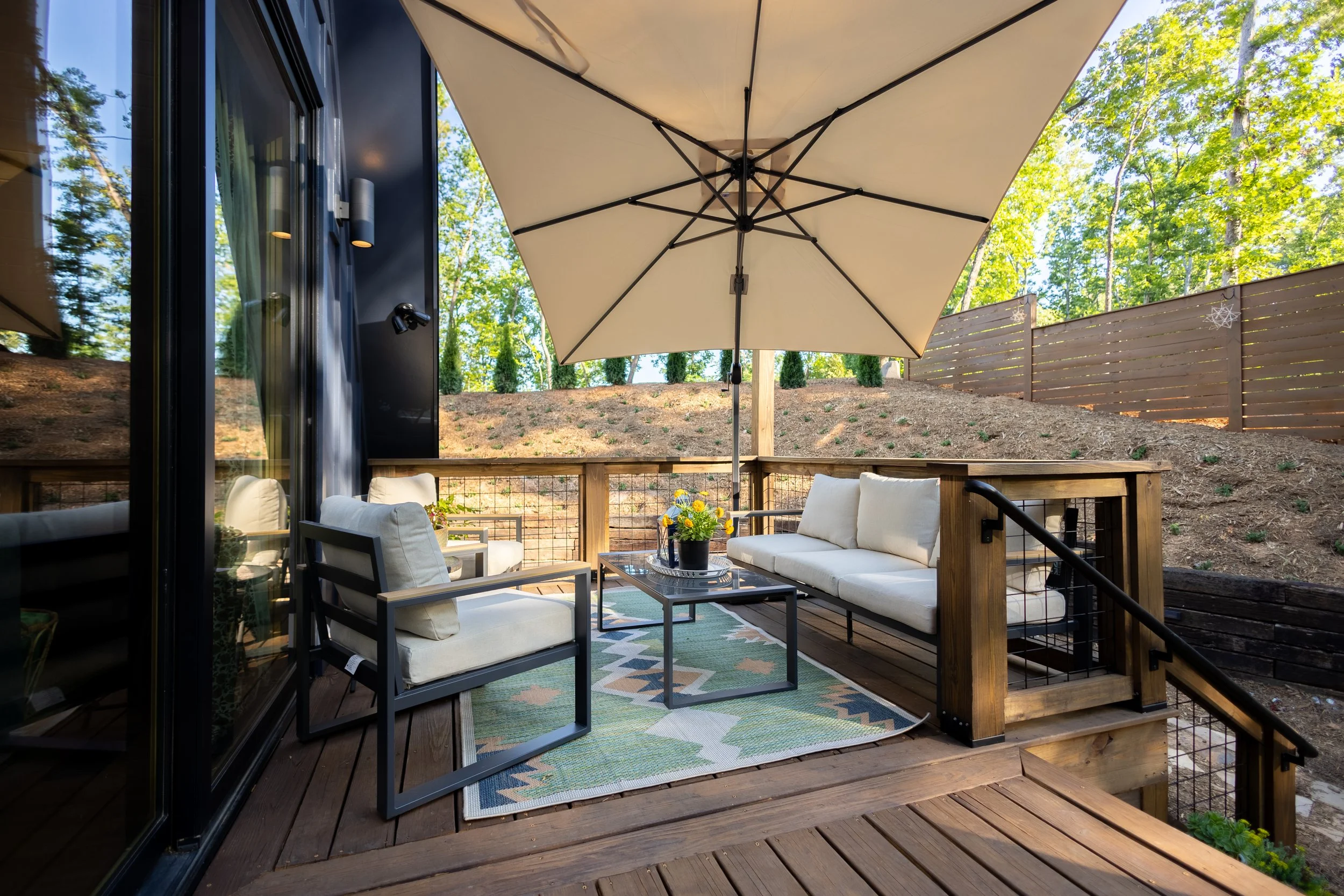 Outdoor balcony with white cushioned seating, a glass-topped table with a potted plant, a large beige umbrella, and a view of green trees and a wooden fence.
