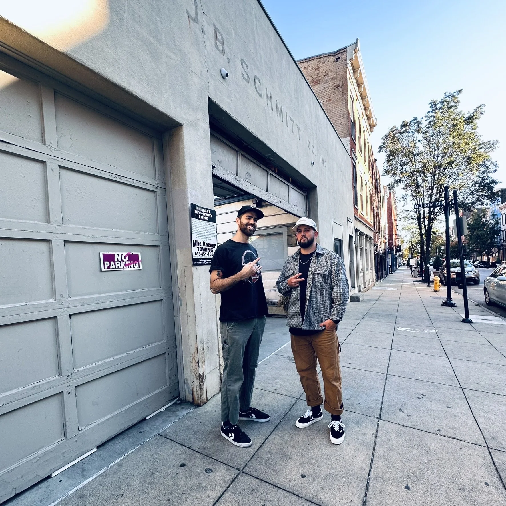 Two men standing outside near a garage with a sign that reads 'NO PARKING' and another sign with contact info for a towing company. They are smiling and posing for the photo, with trees and buildings in the background on a city sidewalk.