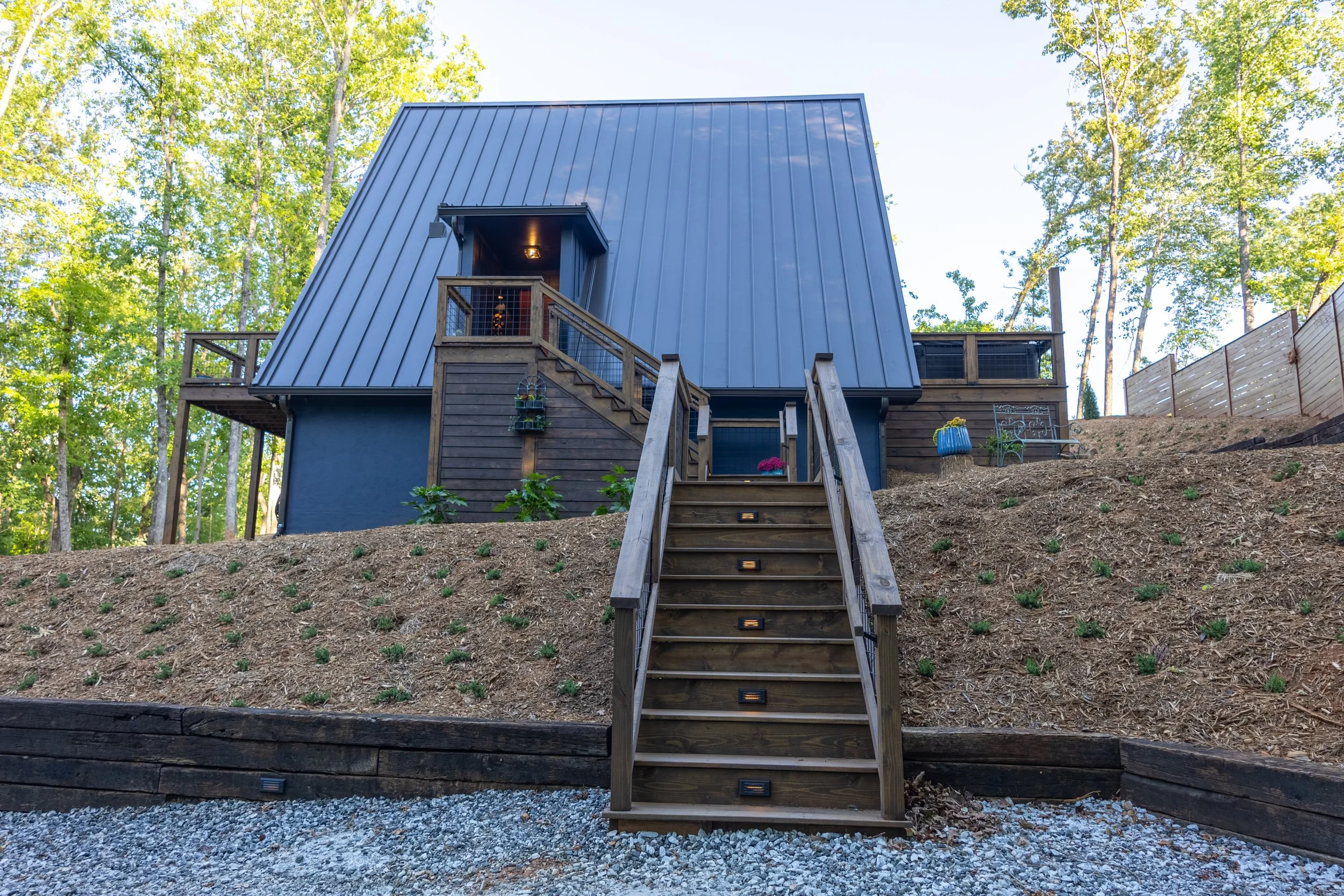 A house with a steep, metal roof and wooden stairs leading up to a small balcony entrance, surrounded by trees and a wooden fence.