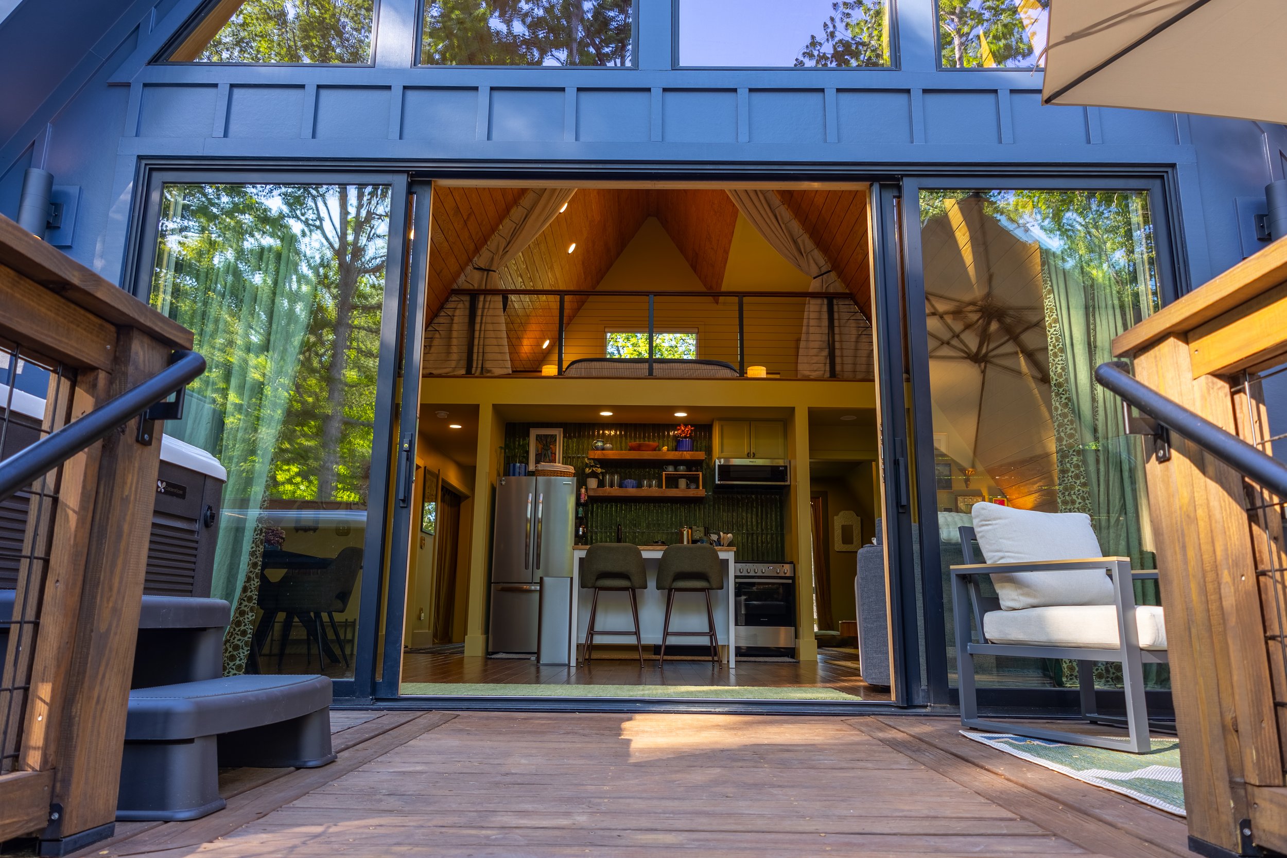 View of a modern house interior from a wooden deck, with large sliding glass doors opening to a dining and living area with a vaulted wooden ceiling, kitchen appliances, and outdoor furniture with an umbrella and trees reflected in the windows.
