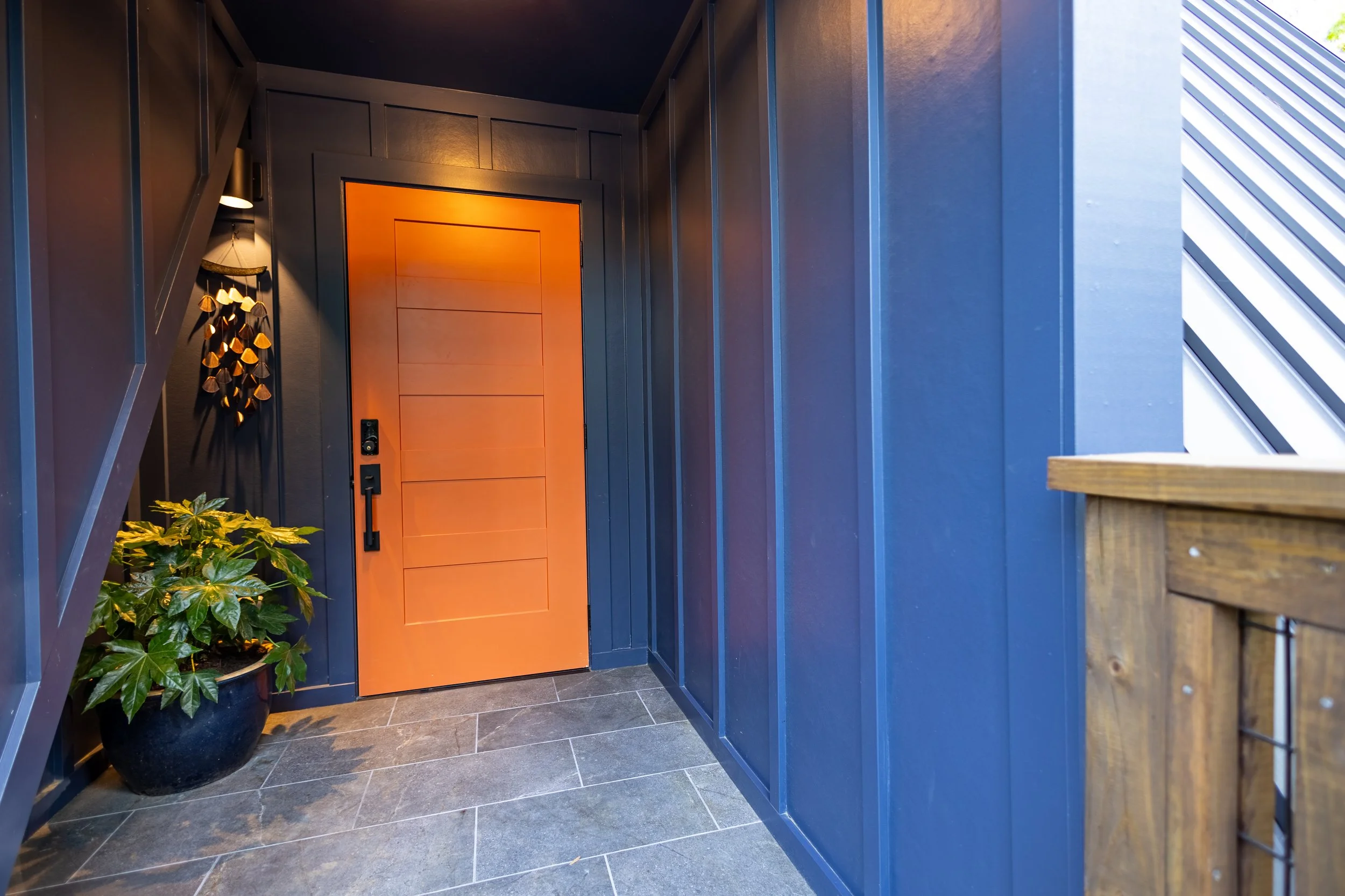 An interior view of a modern hallway with a vibrant orange door set in dark blue walls. There are decorative elements including a plant in a large black pot and a hanging wall decor piece with small mushroom shapes. The floor is tiled with dark gray 