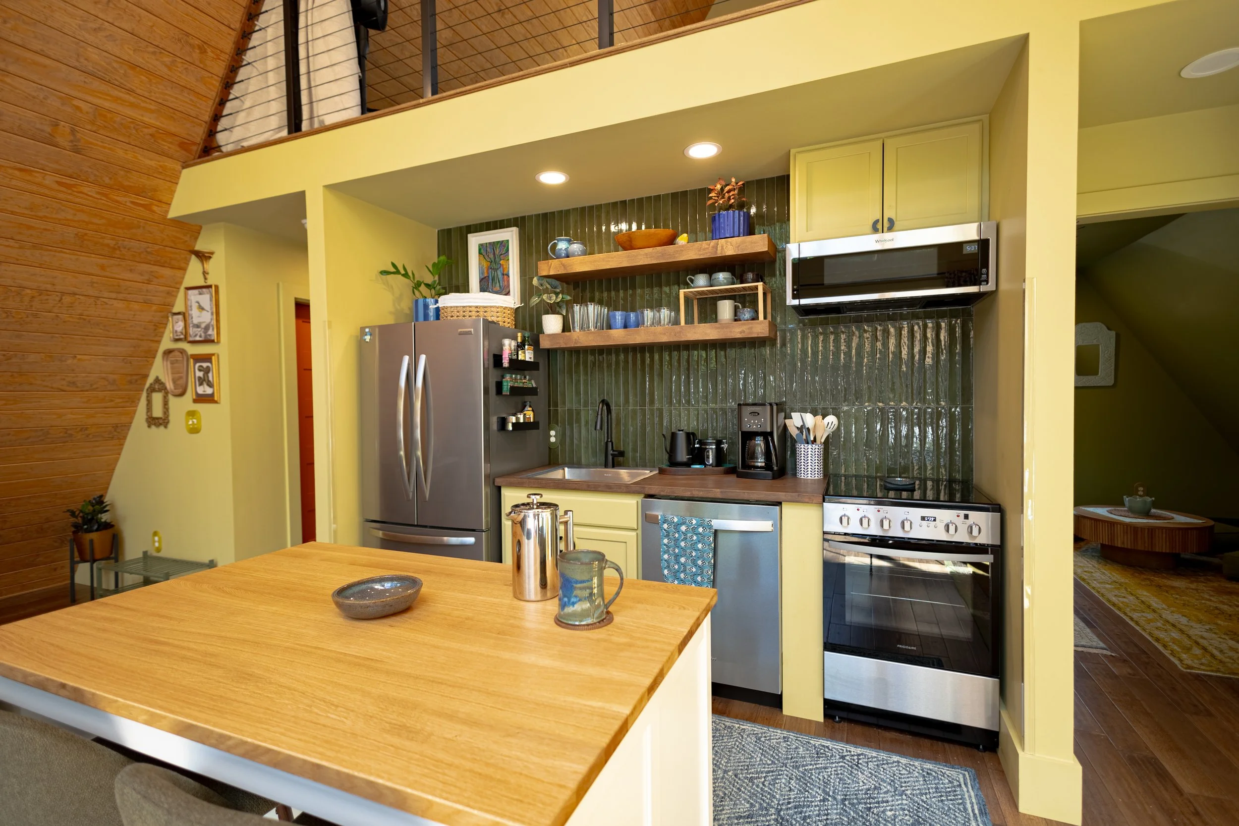 A kitchen in a house with yellow walls, dark green tiled backsplash, open wooden shelves holding dishes and decorative items, stainless steel fridge, microwave, black coffee maker, and stove, in a room with wooden flooring and a wall with picture fra