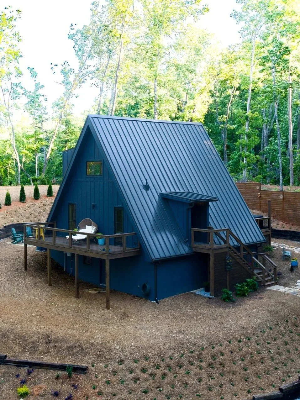 A small, blue A-frame house with a metal roof, surrounded by a wooded backyard with a deck and outdoor seating.