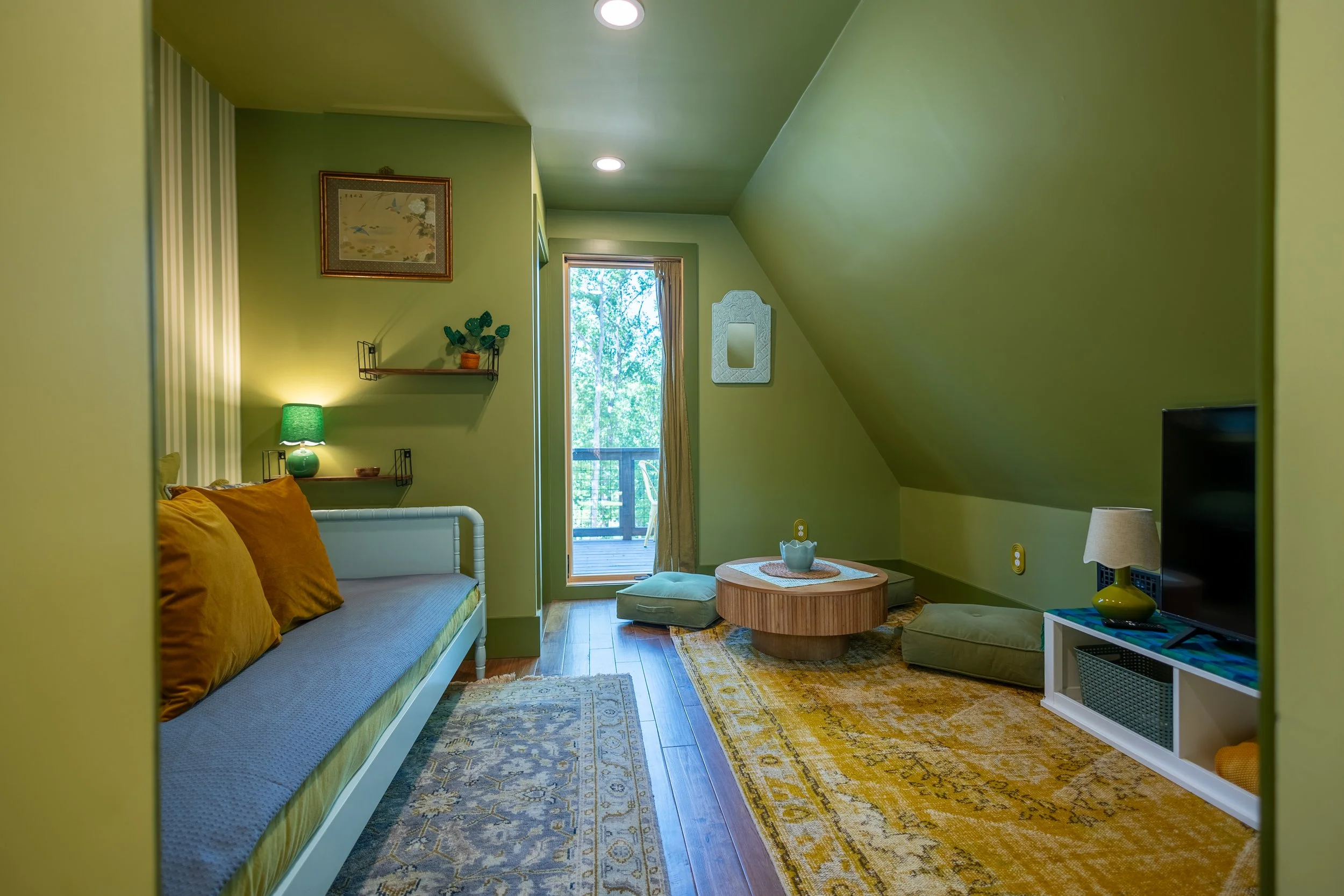 A cozy living room with green walls, a daybed with orange pillows, a small round coffee table, a window leading to a balcony, and various decorative items including a lamp, framed artwork, and shelves.