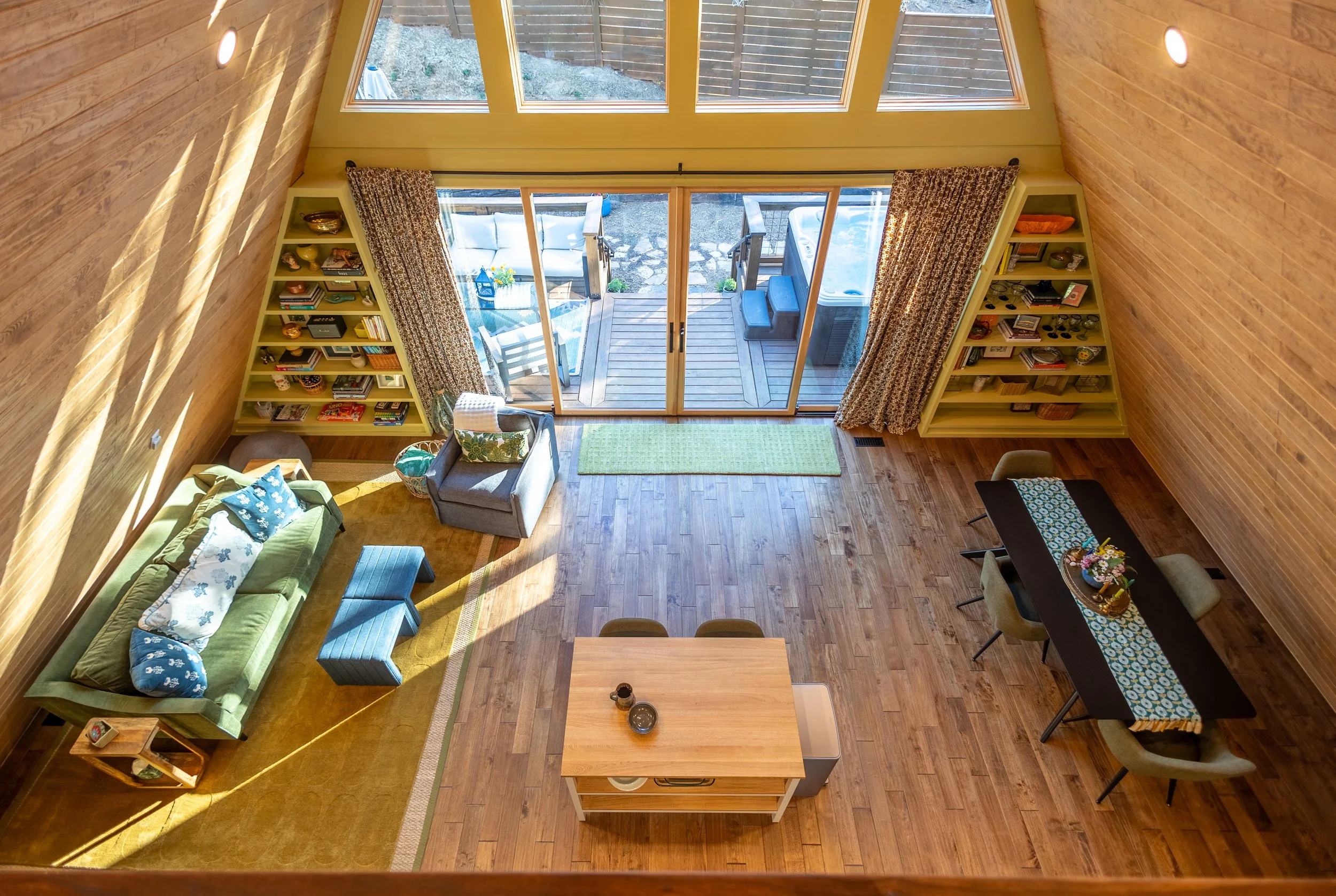 Living room with wooden walls, large glass sliding doors leading to a wooden deck outside with outdoor furniture, bookshelves on either side, a green sofa, a gray armchair, and a wooden coffee table.