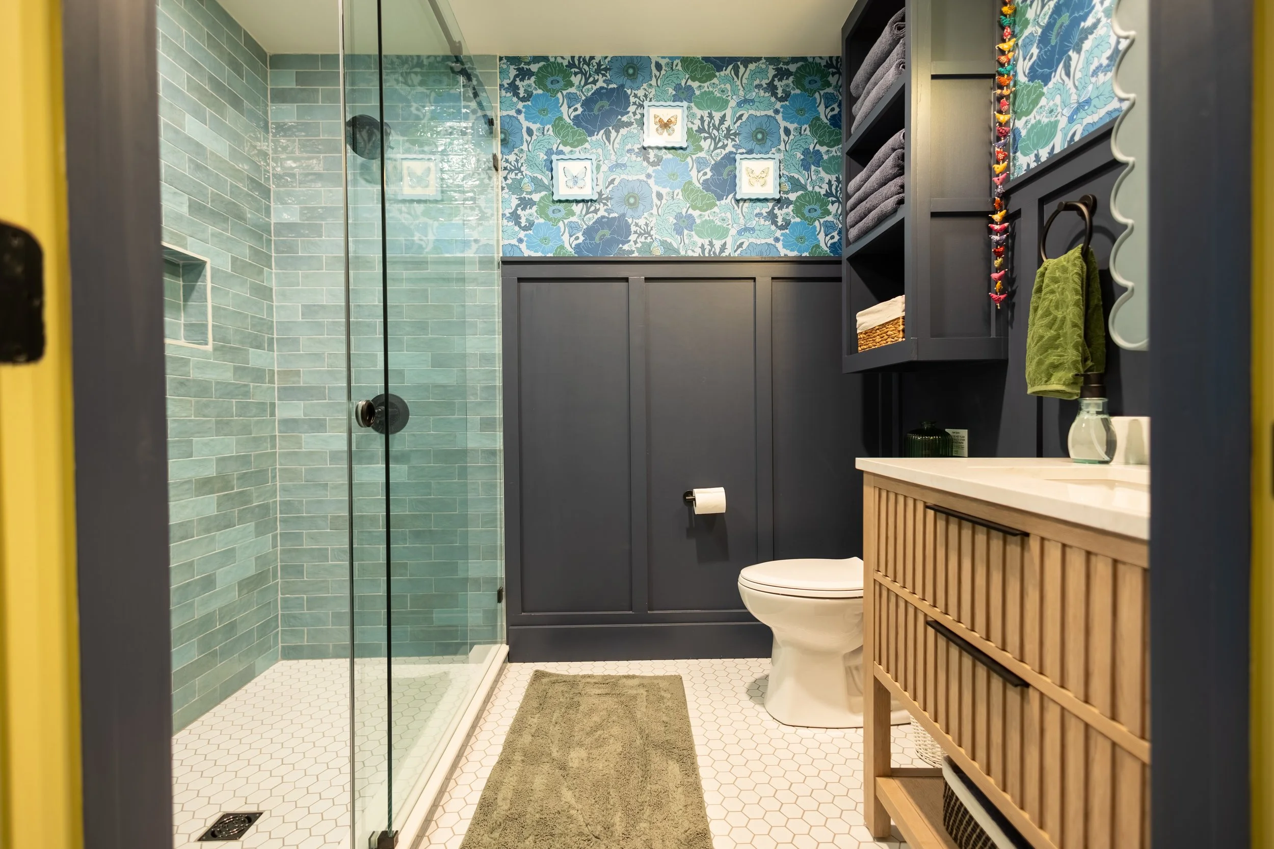 A bathroom with a glass shower, dark cabinetry, a toilet, a wooden vanity, and decorative wallpaper with blue and green floral patterns.