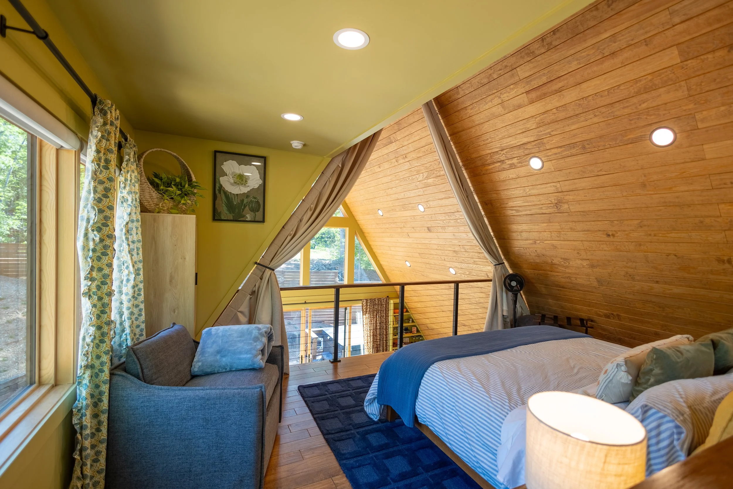 Interior of a cozy bedroom with wooden sloped ceiling, yellow wall, large windows with patterned curtains, a bed with a blue blanket, a gray armchair with a blue cushion, a framed floral artwork, and a balcony view of greenery outside.