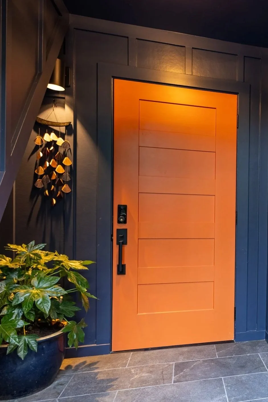 Entrance door with a natural wood finish, black smart lock, and black handle, surrounded by dark gray framing, with a potted green plant to the left and decorative wooden wind chimes hanging on the wall, illuminated by warm lighting.