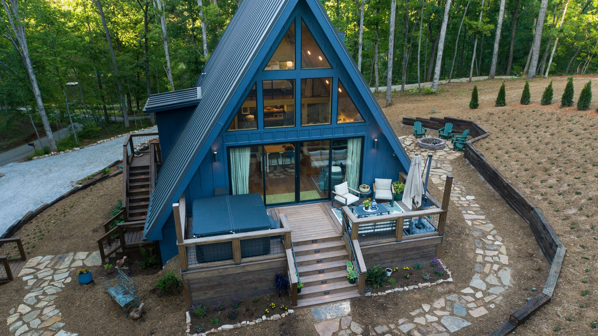 A modern A-frame cabin with large glass windows, blue exterior, and wooden deck with outdoor furniture, surrounded by trees and a landscaped yard with a fire pit and Adirondack chairs.