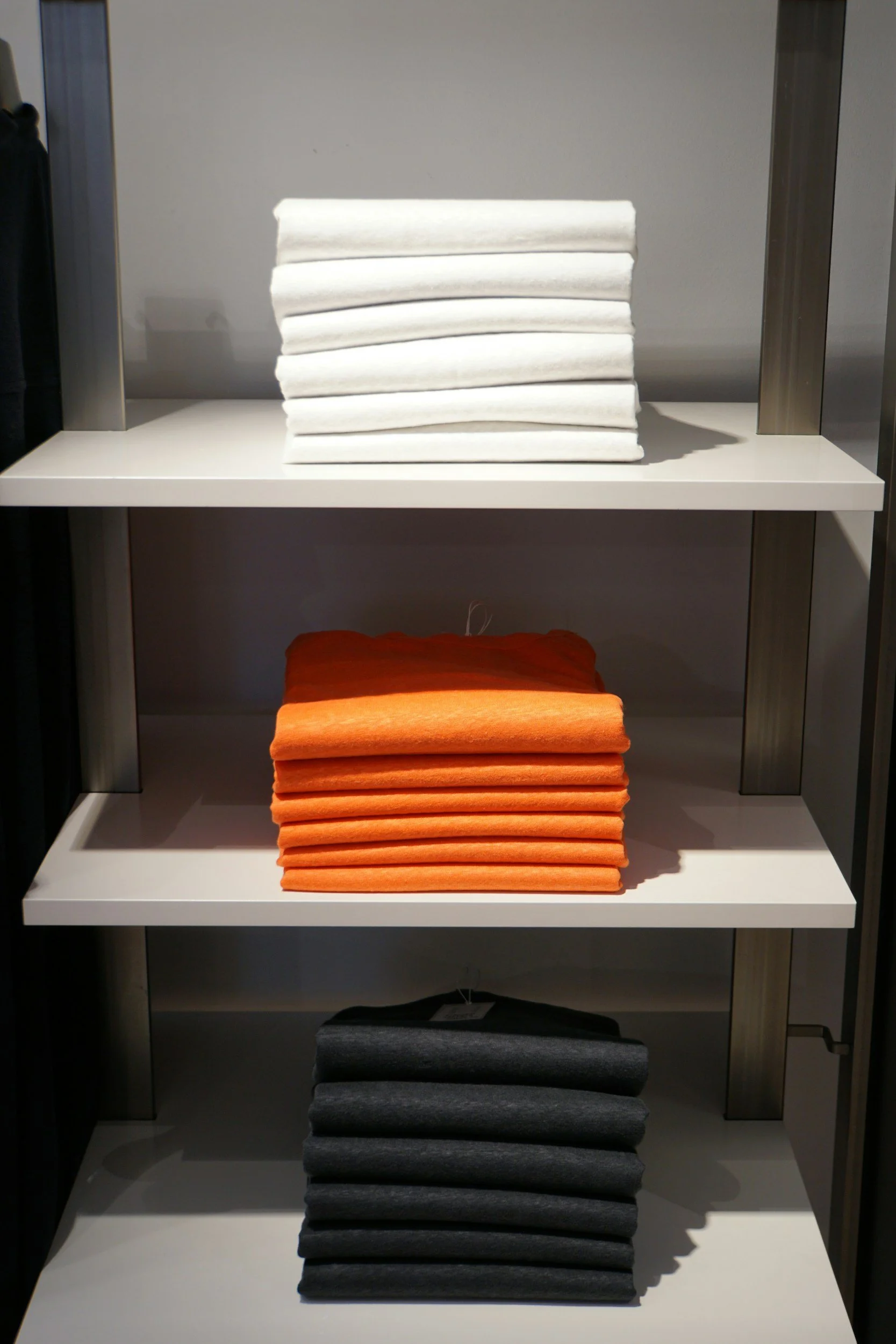 Stacks of folded T-shirts on retail shelves, with white T-shirts on top, orange in the middle, and dark gray at the bottom.