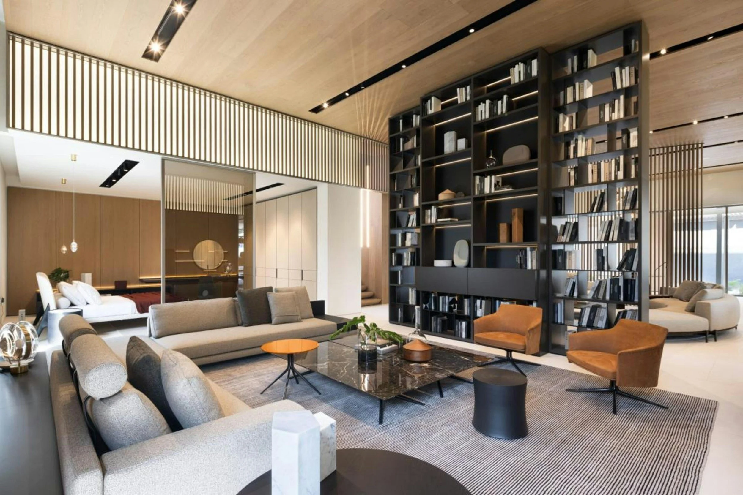Modern living room with beige sofas, a black granite coffee table, and two brown armchairs. A large black bookshelf filled with books separates the room. The space features wood paneling on the ceiling and walls, with built-in lighting and a window allowing natural light.