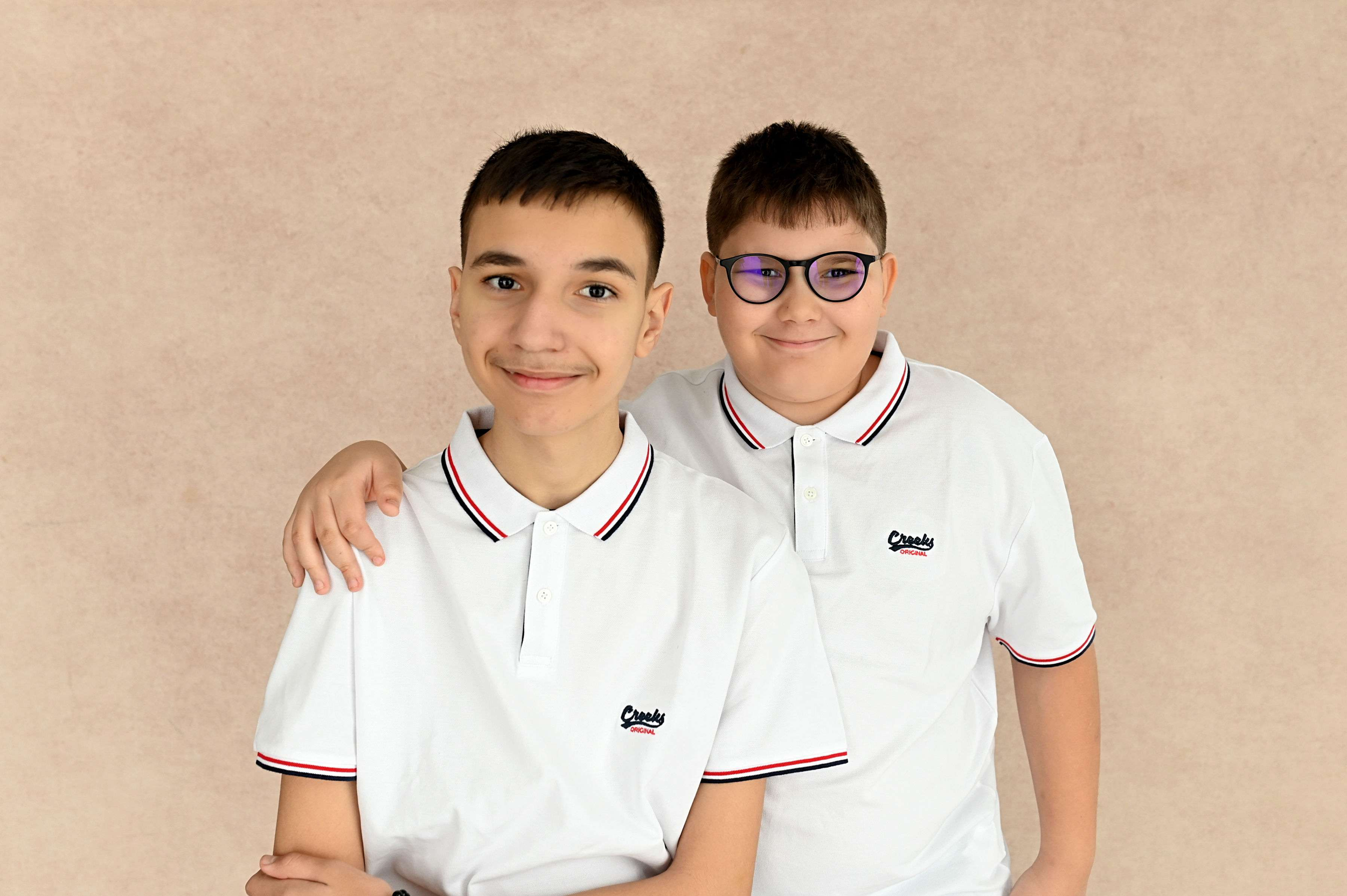 Deux jeunes garçons en chemises blanches avec col orné de rayures rouges, noires et blanches, souriants, posant ensemble contre un mur beige, l'un avec des lunettes