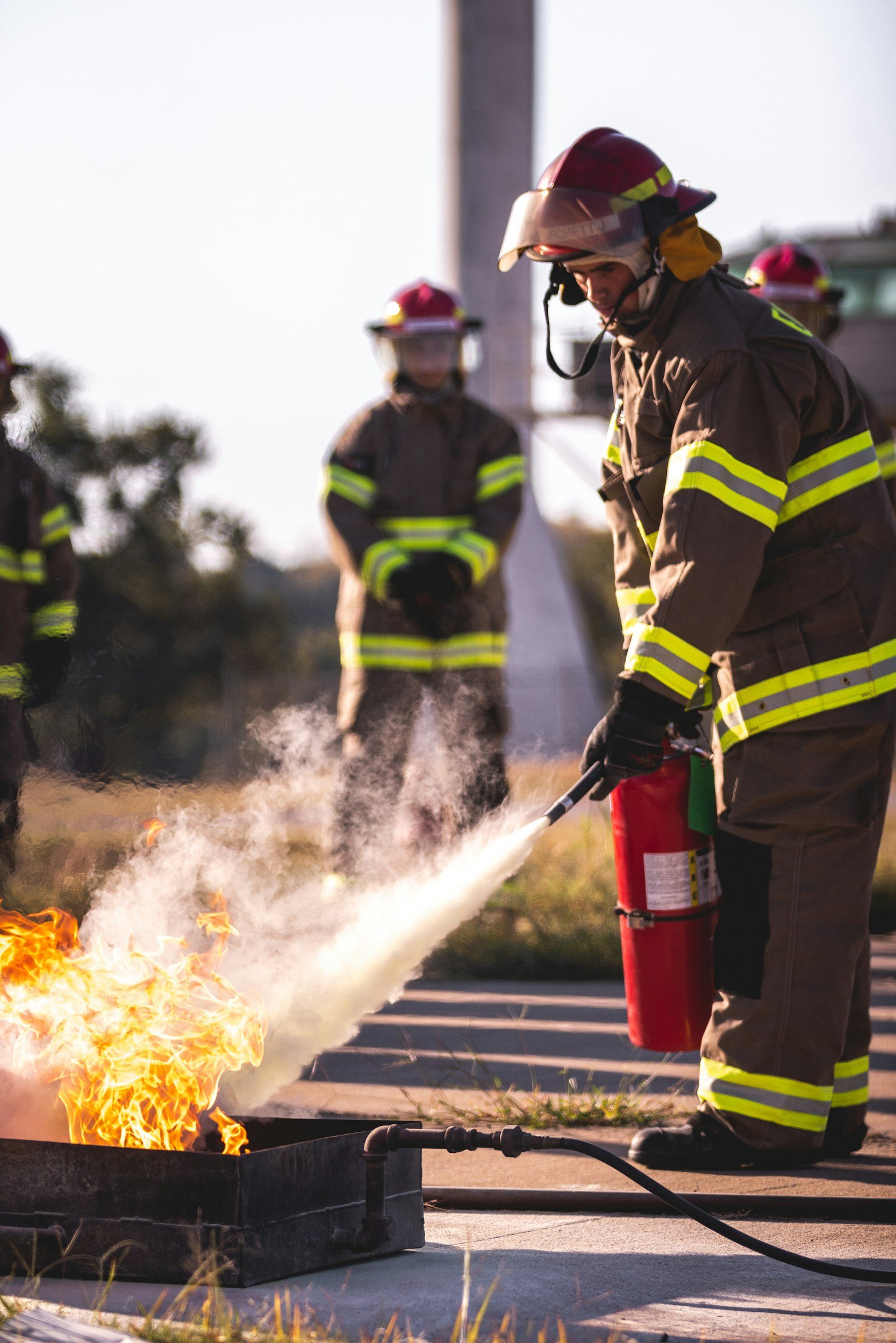 Firefighters wearing protective gear and helmets working to extinguish a small fire with a fire extinguisher outdoors during daylight.