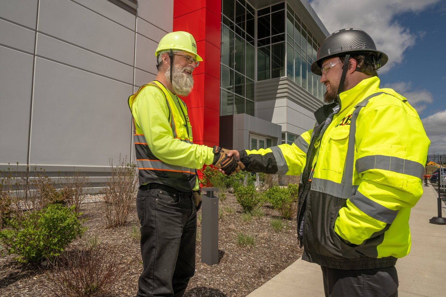 Two construction workers shaking hands outside near a modern building, both wearing yellow safety vests and helmets.