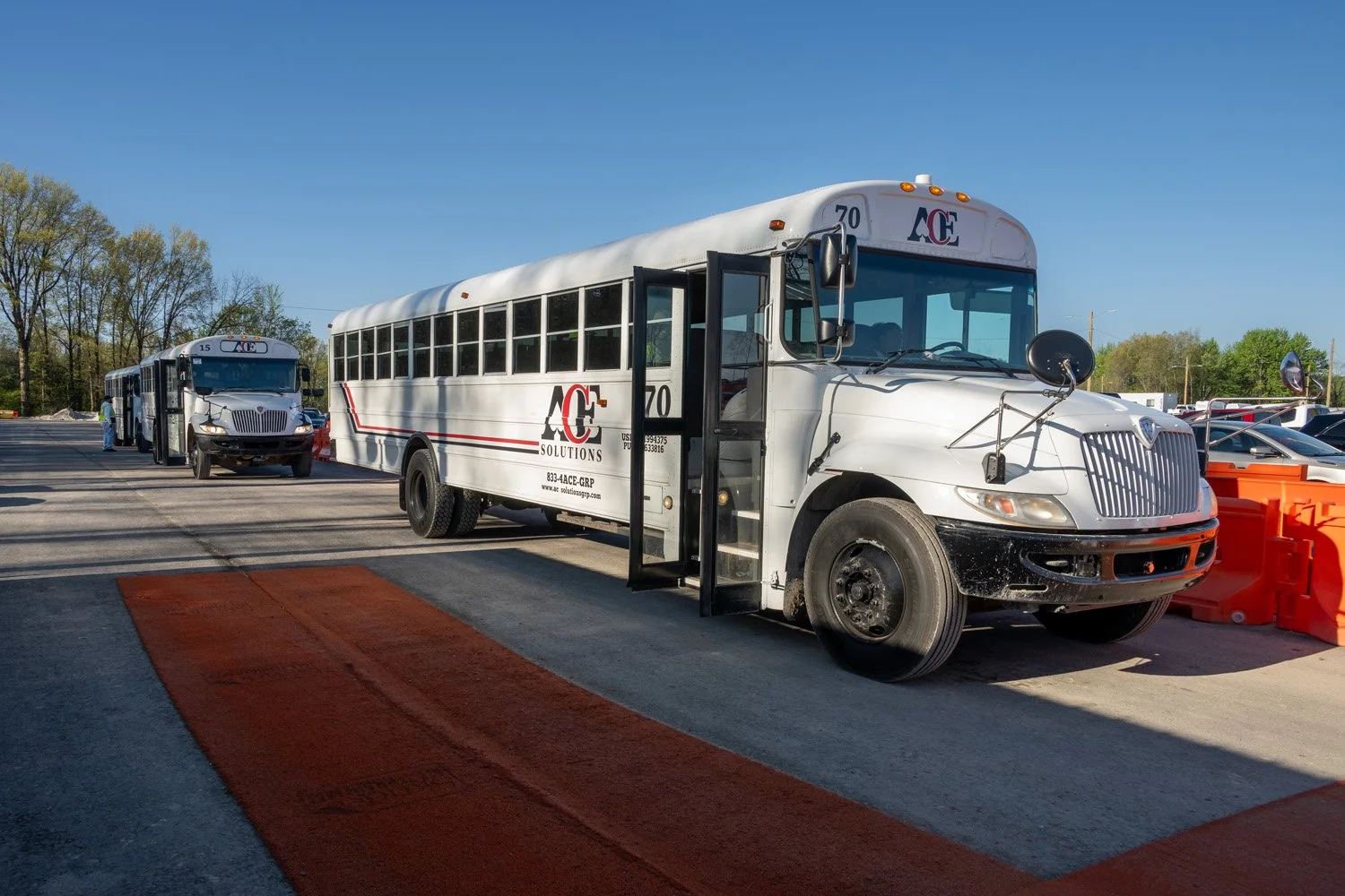 A row of shuttle buses parked outdoors under a clear blue sky, with trees in the background and orange barriers in the foreground.
