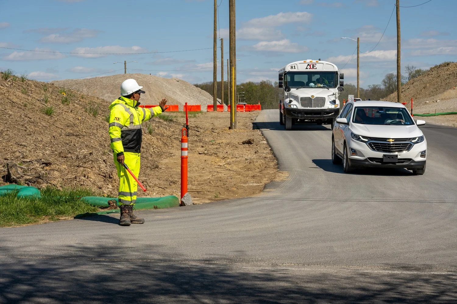 A construction worker wearing a yellow high-visibility safety suit and white helmet standing on the side of a road, signaling to vehicles to stop or slow down. A white truck and a silver SUV are driving towards the worker, with construction barriers and utility poles in the background under a partly cloudy sky.