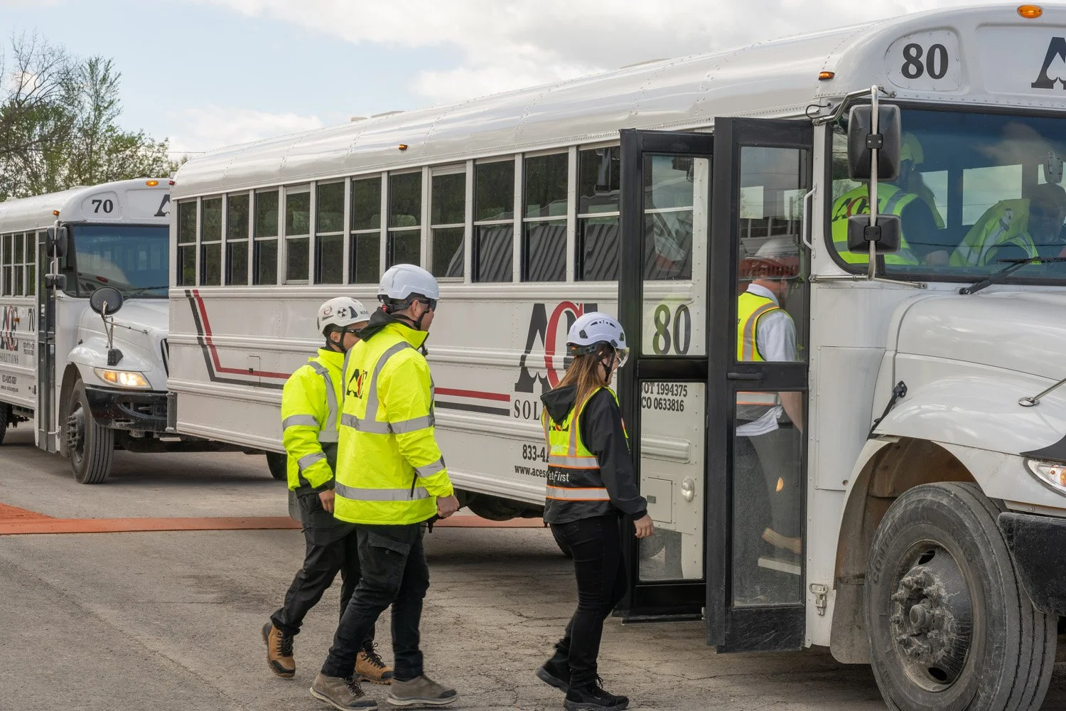 Group of people wearing safety vests and helmets walking past a school bus with a sign that says 'ACE SOLUTIONS' on it.