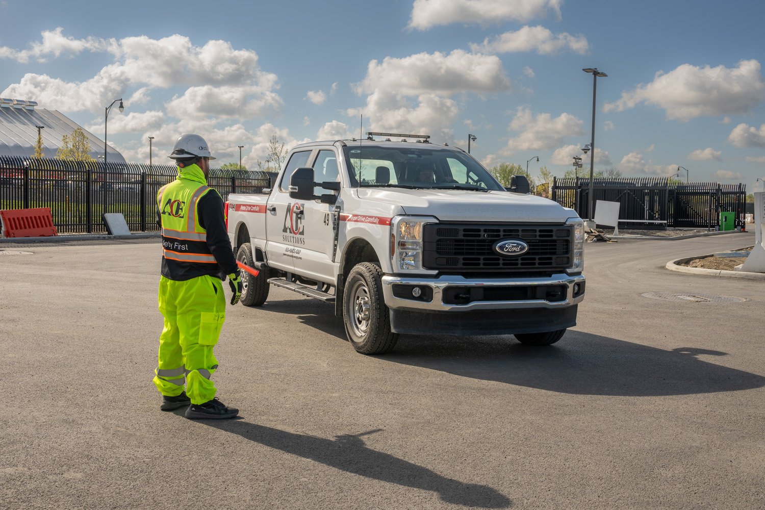 Construction worker in high-visibility clothing and safety helmet directing traffic near a white Ford pickup truck at a construction site on a sunny day.
