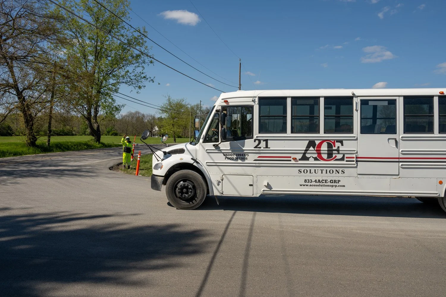 A white bus with the logo of ACE Solutions parked on a quiet rural road, with a construction worker in a neon yellow safety suit and helmet standing nearby holding a traffic cone, surrounded by green trees and a clear blue sky.