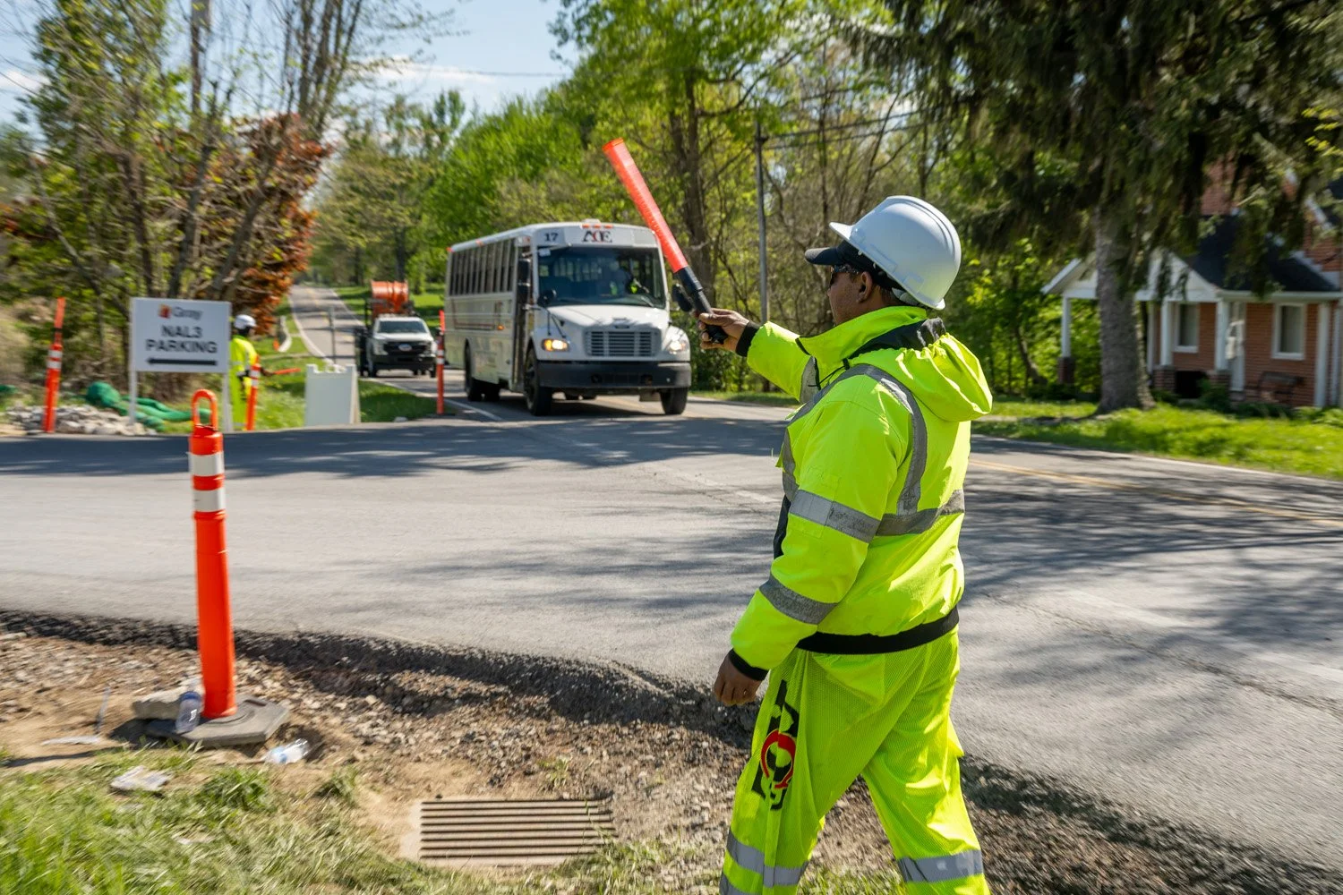 A construction worker wearing a yellow high-visibility safety jacket and helmet, directing traffic with a red baton on a residential street with trees, houses, and a bus approaching.