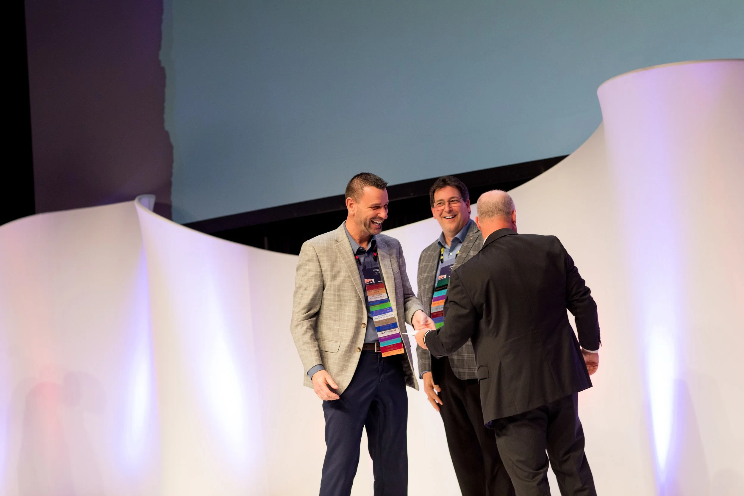 Three men standing and talking at a conference or event, with one man shaking hands with another. All three are smiling and appear to be engaged in conversation. The background has a large screen and white, curving panels.