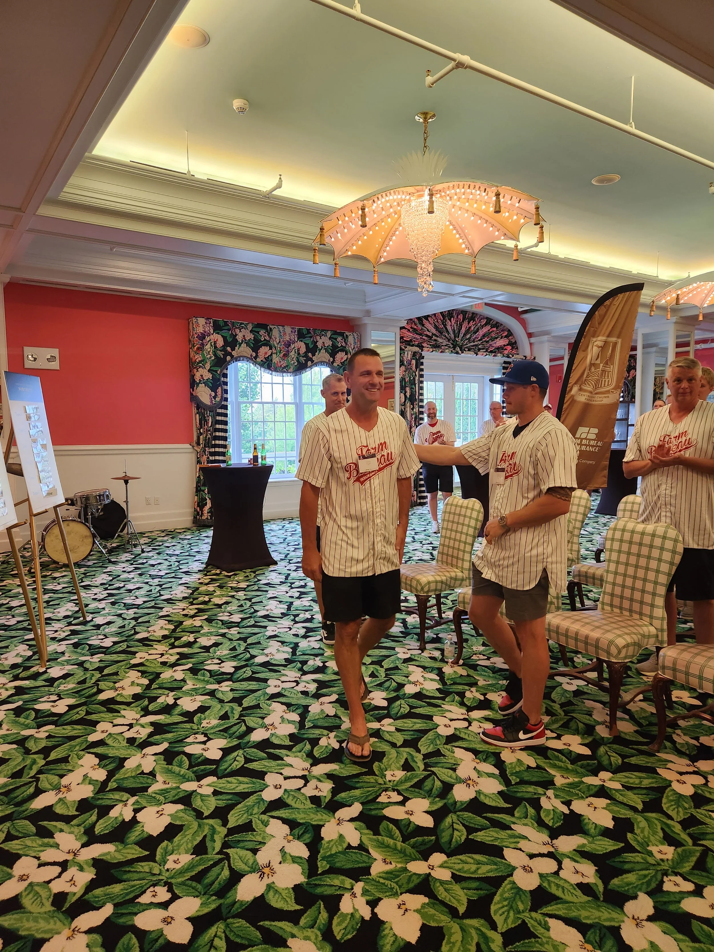 People in baseball jerseys are mingling and smiling in a decorated indoor space with a floral carpet, patterned curtains, and chandeliers.