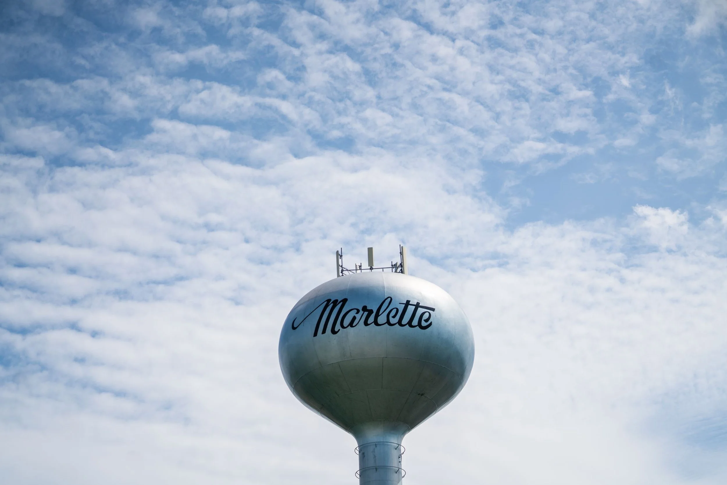 A white spherical water tower with the name "Marlette" written on it, located against a partly cloudy sky with a blue background and some clouds.