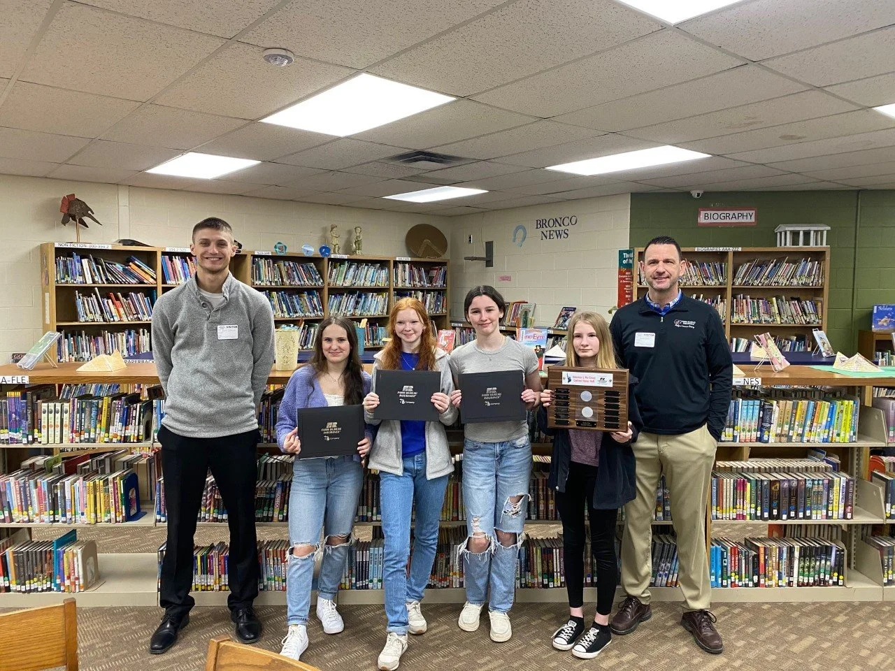 Group of six people, four girls and two men, standing in a school library in front of bookshelves, holding certificates and a trophy, smiling for the camera.