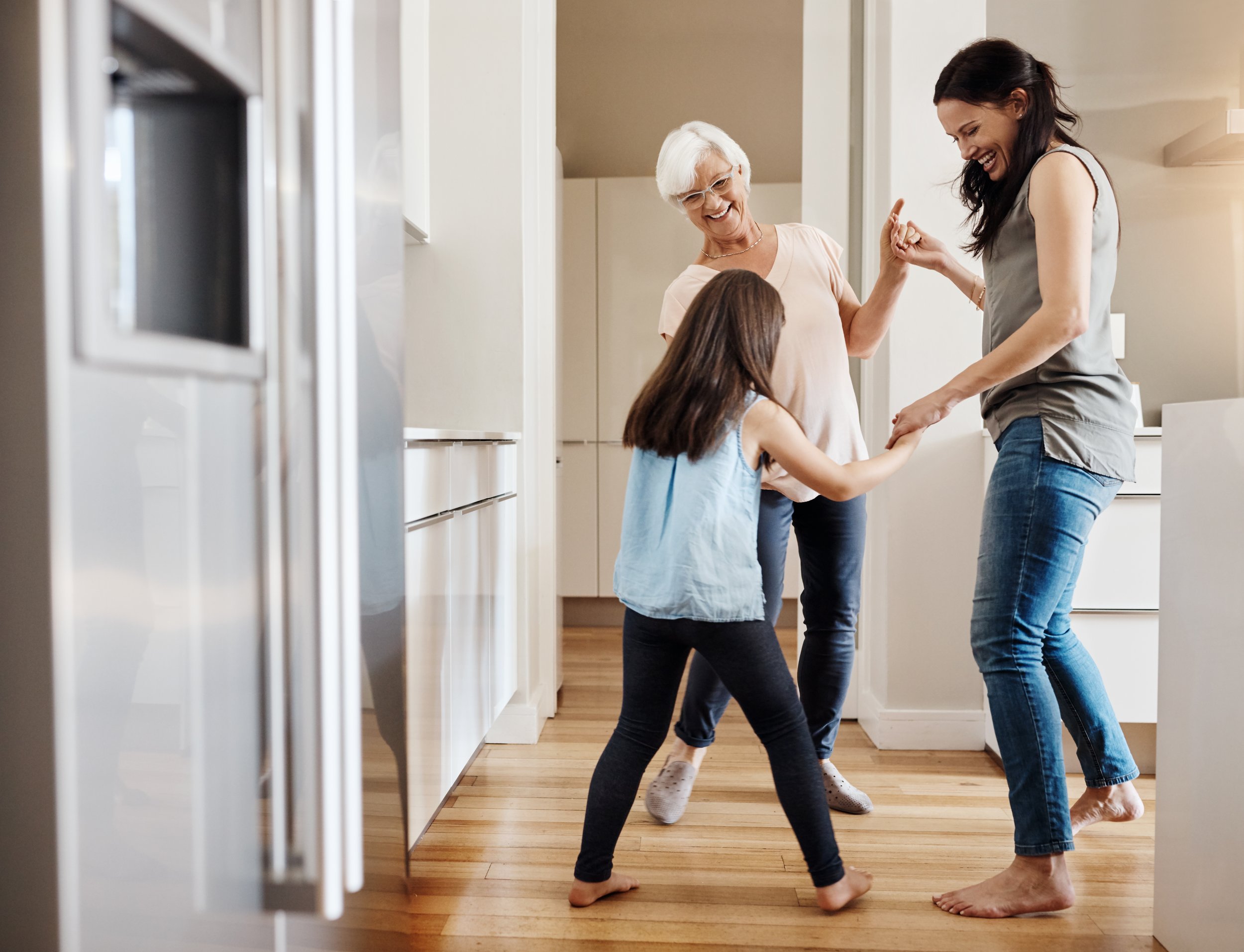 Three generations of women dancing and holding hands in a kitchen with wooden floors, white cabinets, and a refrigerator.