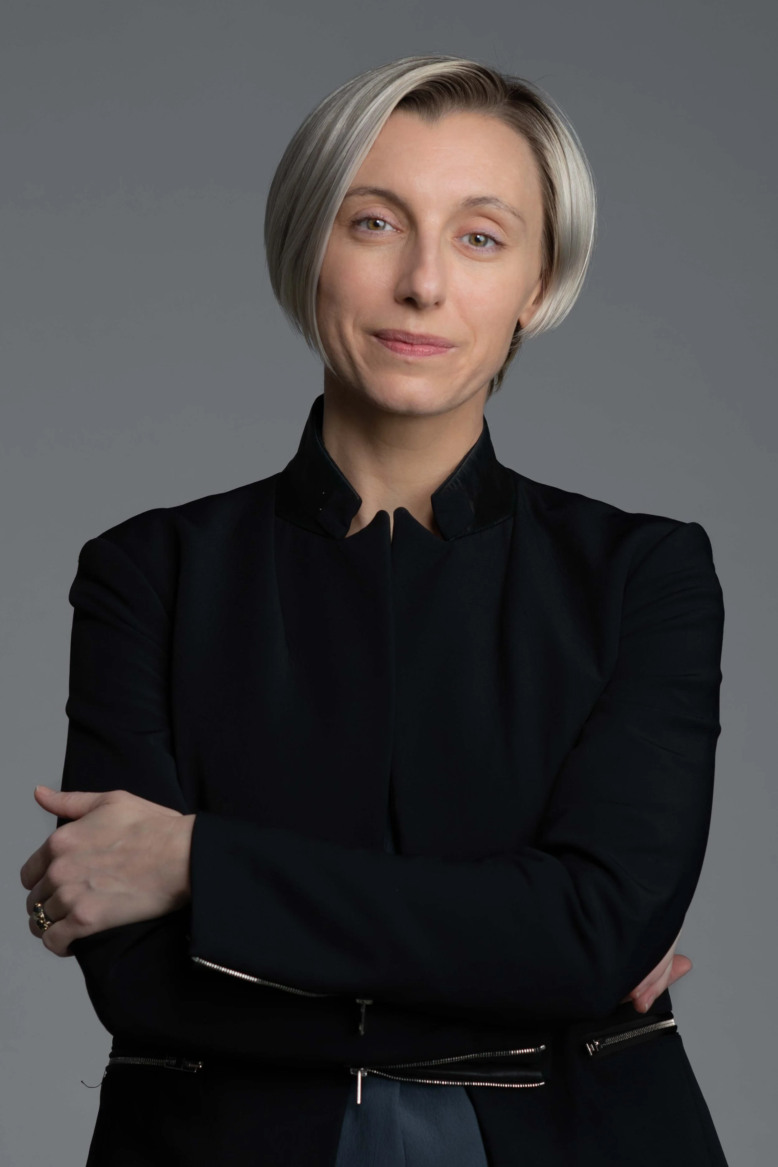 A woman with short blonde hair, wearing a black top and crossing her arms, standing against a gray background.