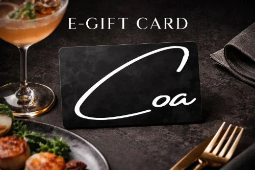 A black E-gift card with white cursive text, placed on a dark table with a cocktail, food, cutlery, and a napkin nearby.
